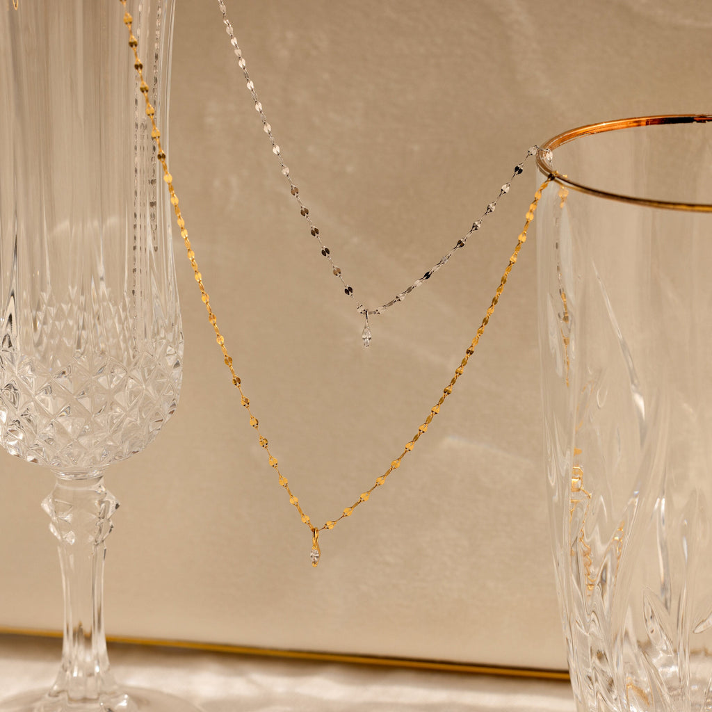 The Dainty Marquise Pendant Necklace is draped over two elegant crystal glasses on a beige background.