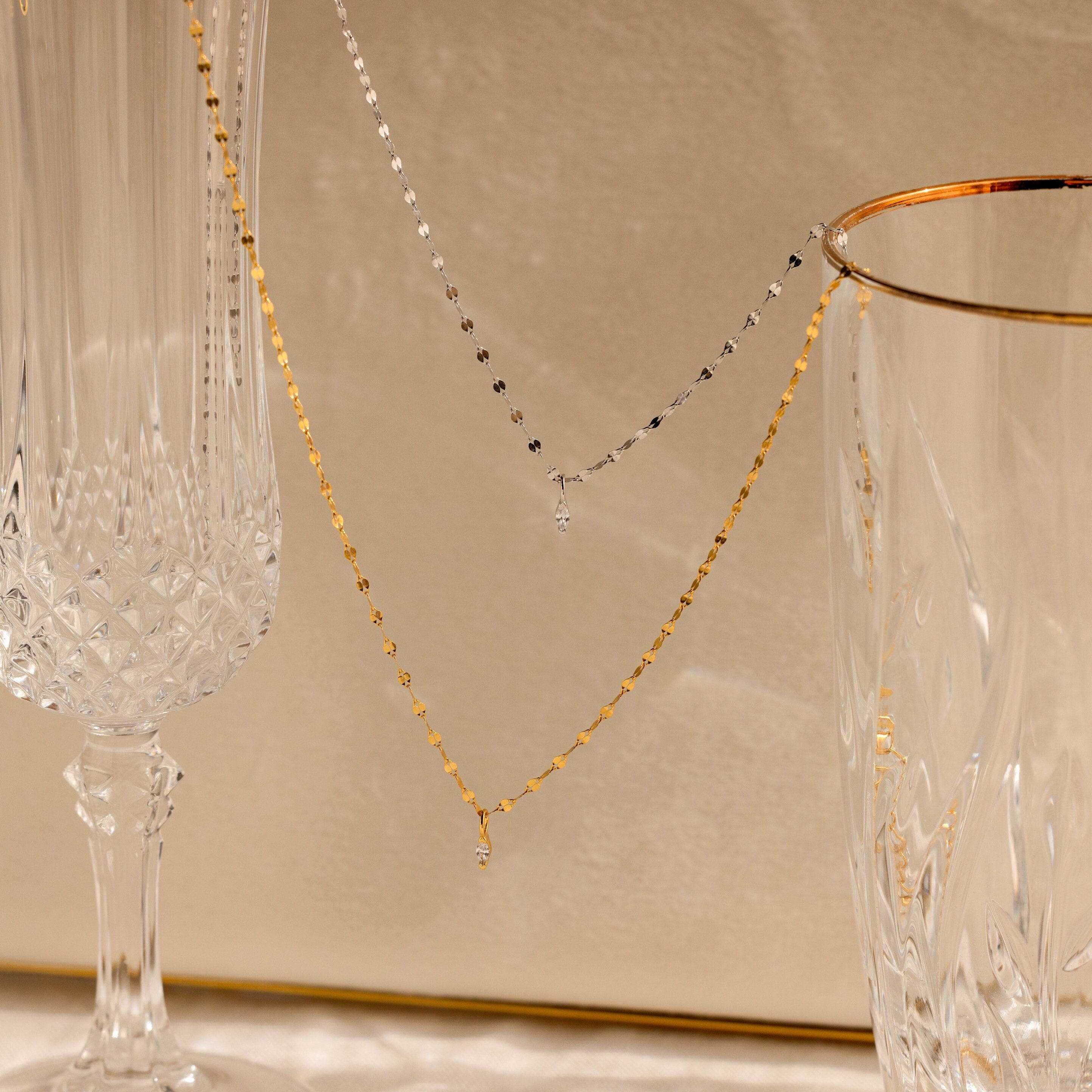 The Dainty Marquise Pendant Necklace is draped over two elegant crystal glasses on a beige background.