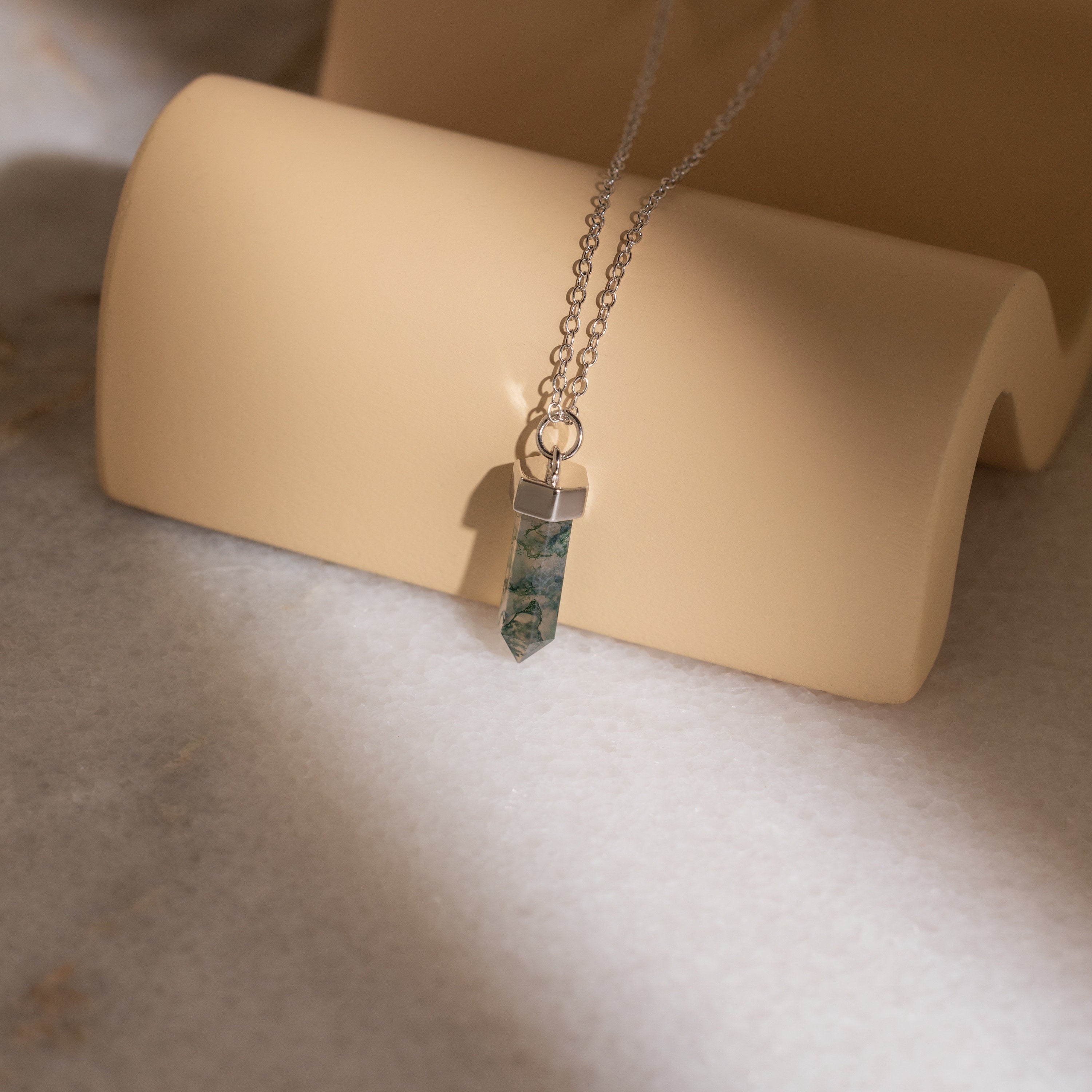 The Agate Quartz Necklace features a marbled green gemstone charm pendant on a silver chain, displayed on a beige curved stand.