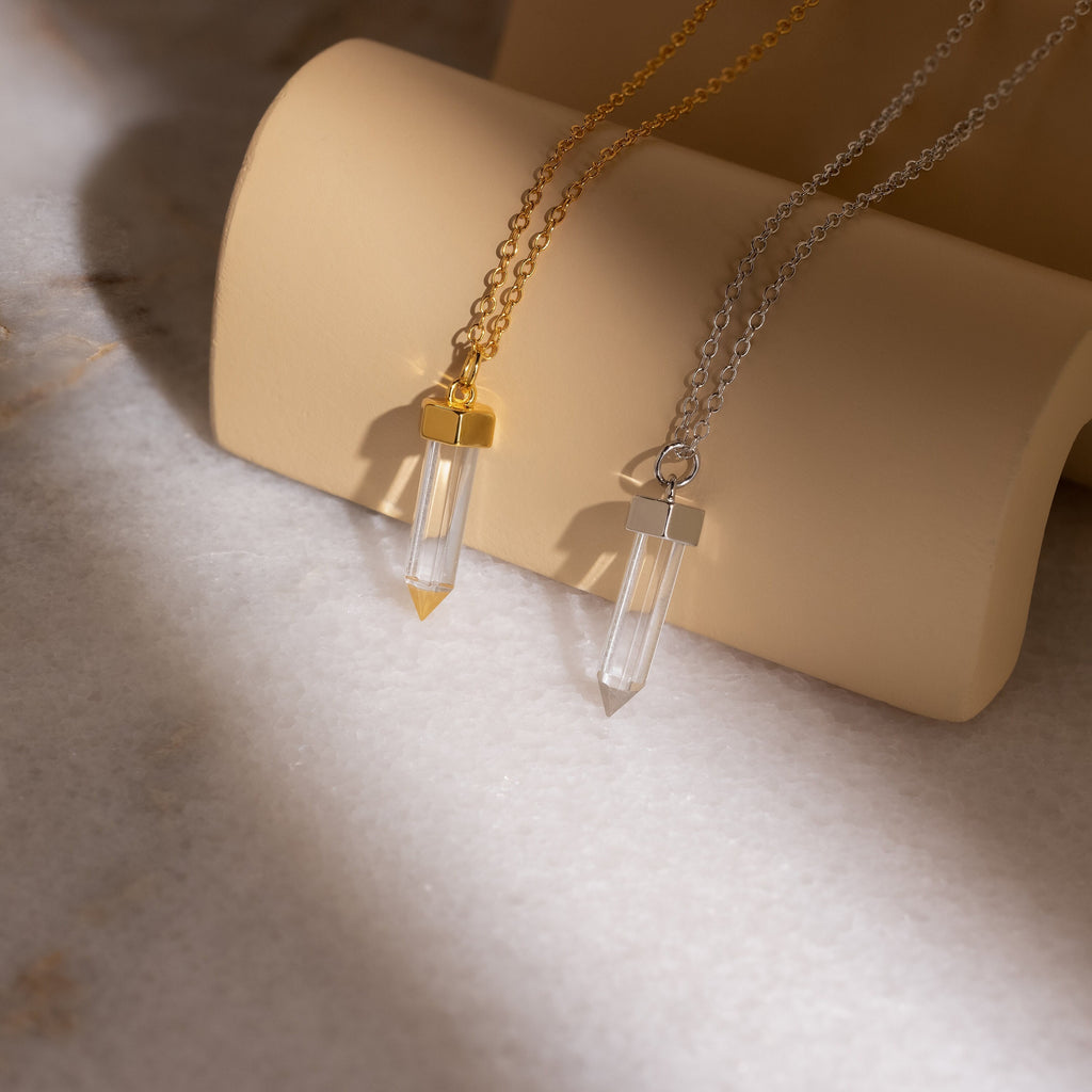 Two Clear Quartz Necklaces, one gold and one silver, featuring hexagonal point pendants, are displayed on a beige curved stand in soft light. Perfect as elegant crystal jewelry or a stunning Clear Quartz Necklace addition.