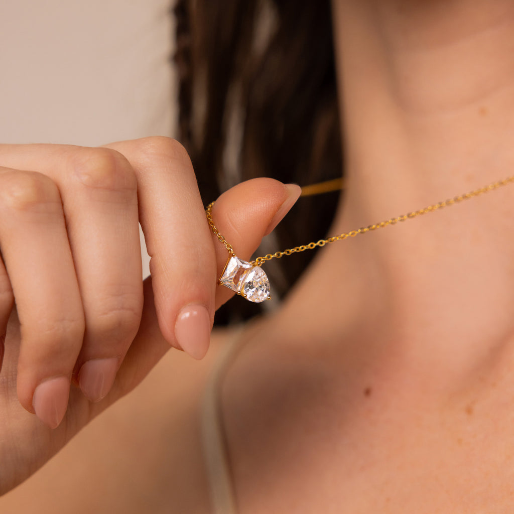 A woman holds the Toi et Moi Diamond Necklace, featuring an elegant teardrop-shaped diamond pendant, between her fingers near her neck.