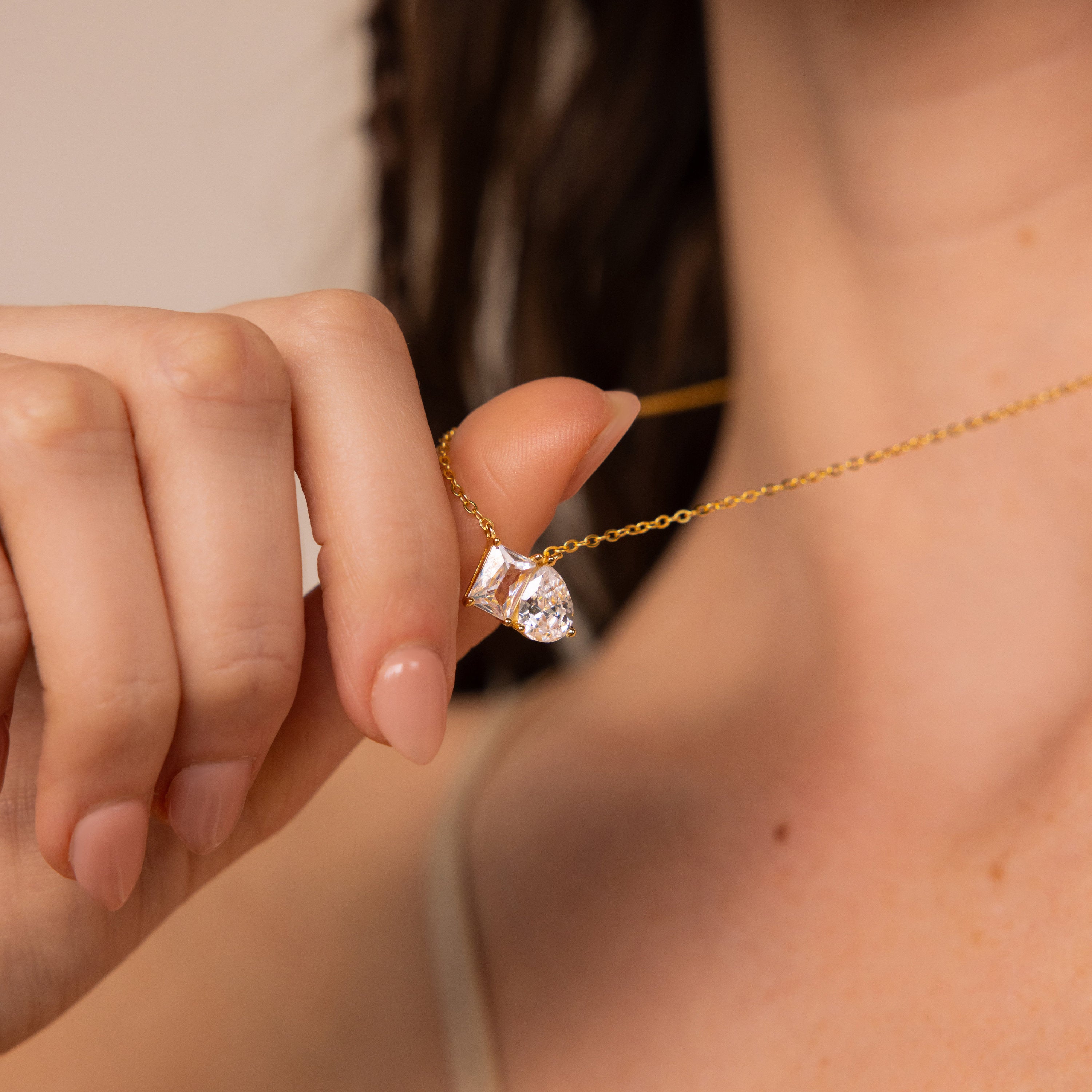 A woman holds the Toi et Moi Diamond Necklace, featuring an elegant teardrop-shaped diamond pendant, between her fingers near her neck.