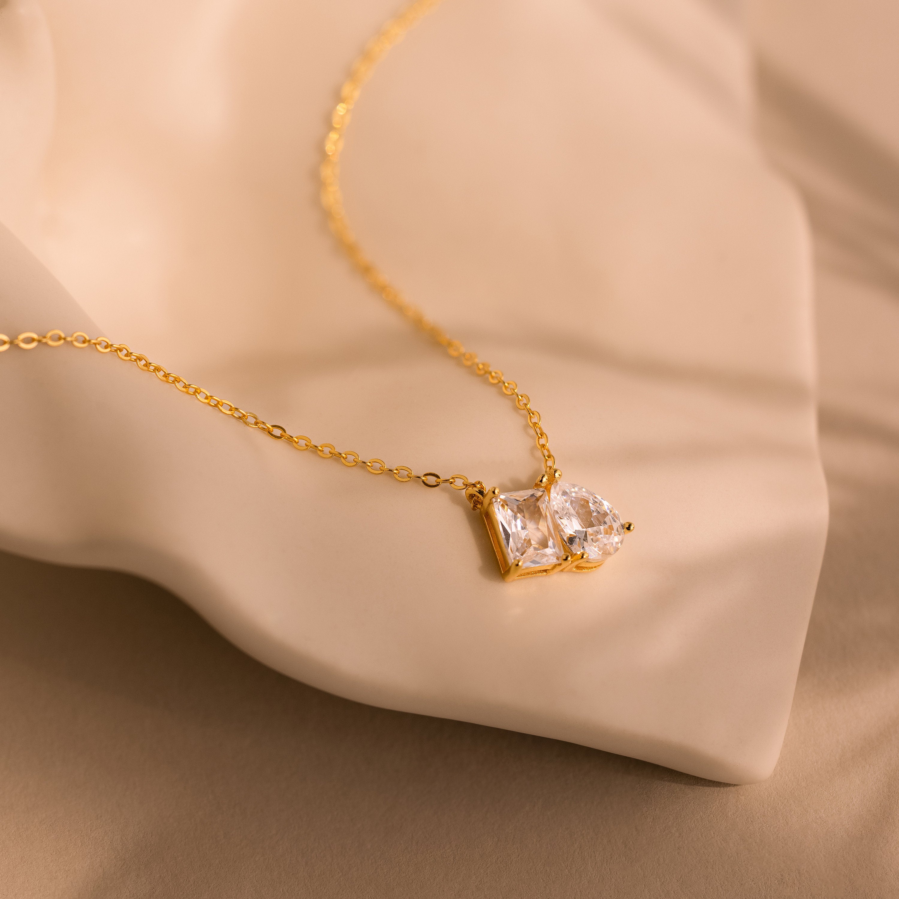Toi et Moi Diamond Necklace featuring an emerald cut diamond pendant sparkling on a soft beige fabric background.