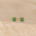 The Emerald Ivy Flower Studs feature small gold floral designs with green gemstones, inspired by nature and ideal for everyday elegance. Displayed on a textured beige surface.