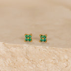 The Emerald Ivy Flower Studs feature small gold floral designs with green gemstones, inspired by nature and ideal for everyday elegance. Displayed on a textured beige surface.