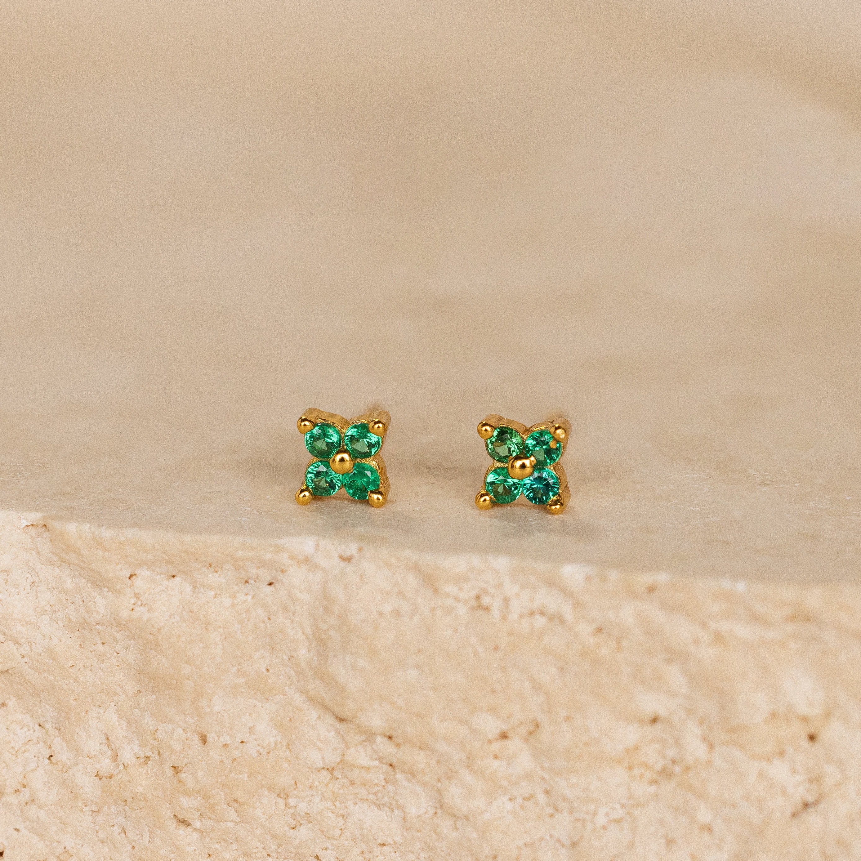 The Emerald Ivy Flower Studs feature small gold floral designs with green gemstones, inspired by nature and ideal for everyday elegance. Displayed on a textured beige surface.
