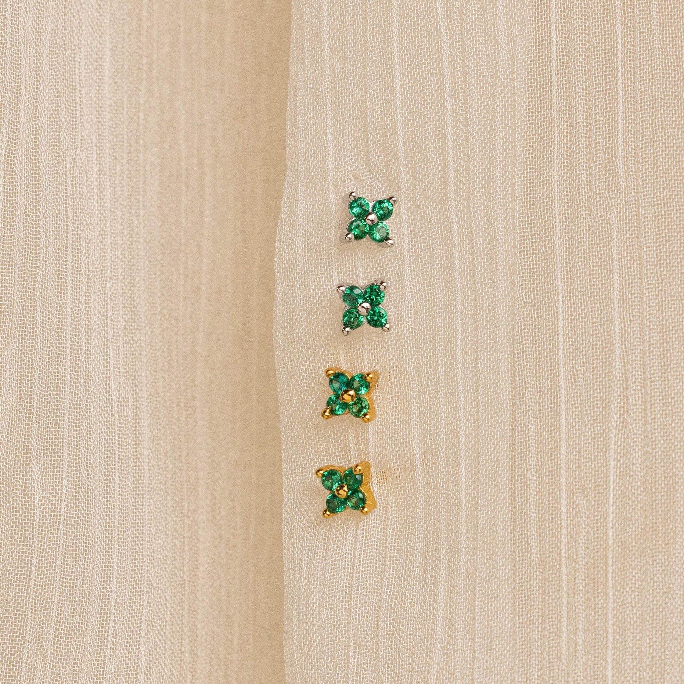 Four pairs of small green gemstone earrings—two silver and two gold—are displayed on beige fabric. The nature-inspired Emerald Ivy Flower Studs add a touch of elegance to any look.