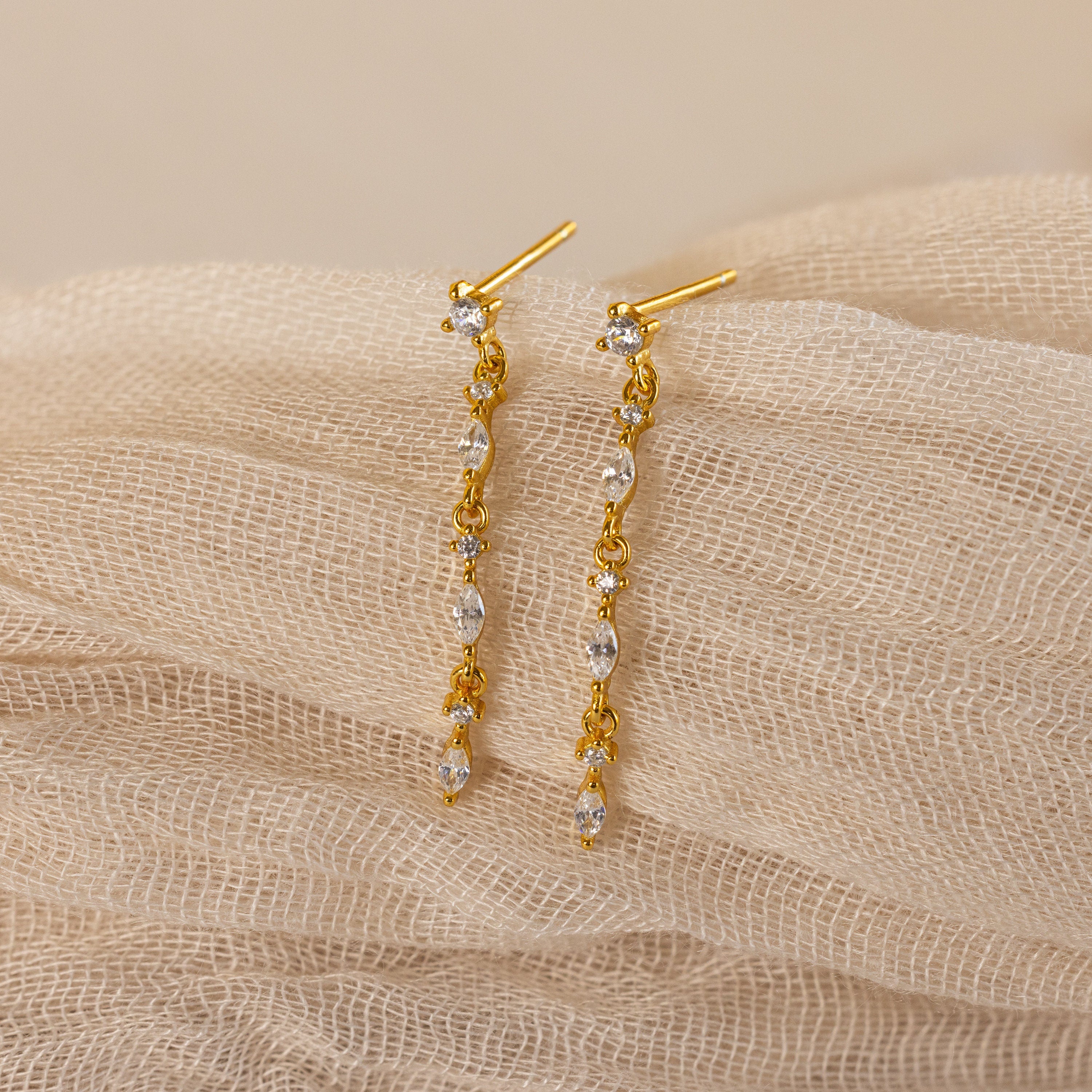 Dainty Diamond Drop Earrings, ideal for weddings or bridesmaids, featuring sparkling crystals and displayed on a soft beige textured fabric background.