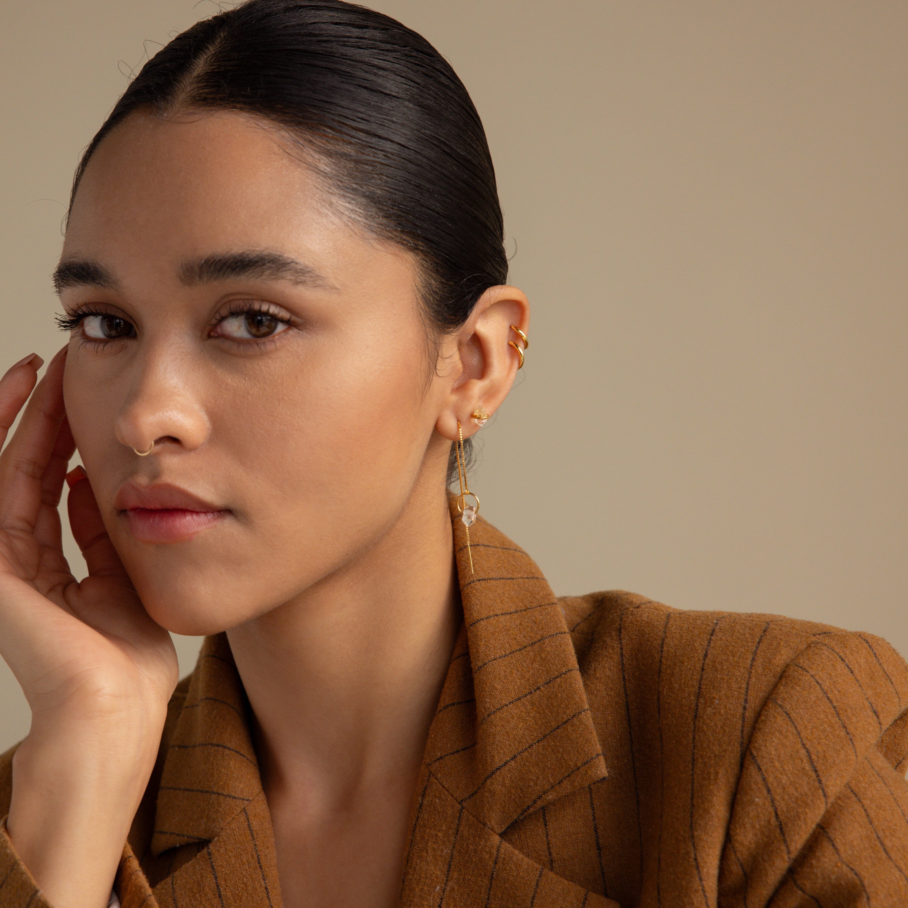 A woman with slicked-back hair wears a brown pinstripe blazer and the Herkimer Diamond Earrings, resting her hand on her face.