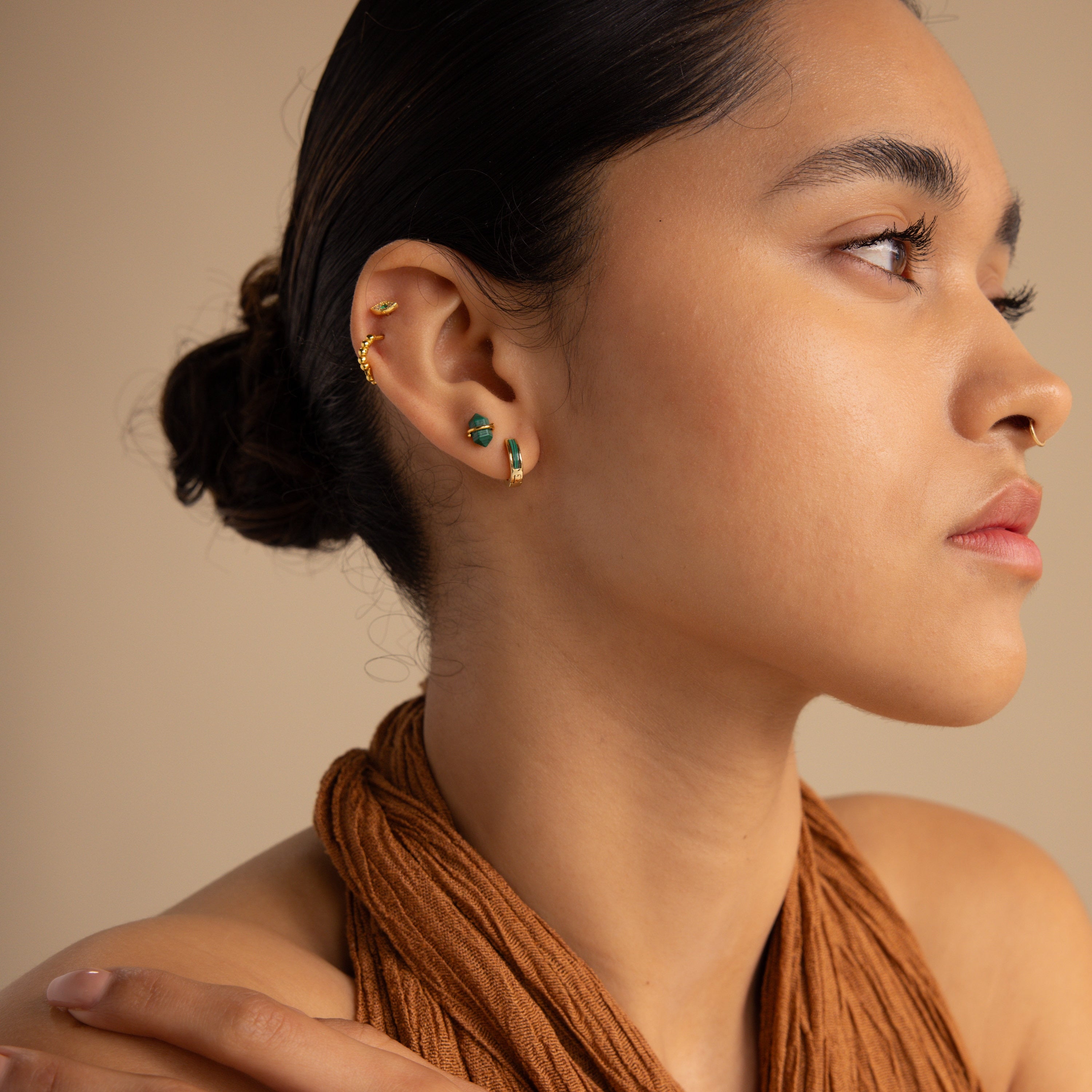 A woman with a scarf around her neck wears Malachite Inlay Huggies, bringing a stylish hint of green stone jewelry to her look.