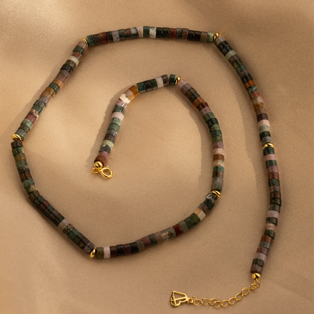 The Moss Agate Beaded Necklace showcases mixed color moss agate beads and gold clasps for earthy elegance, displayed on a beige fabric backdrop.