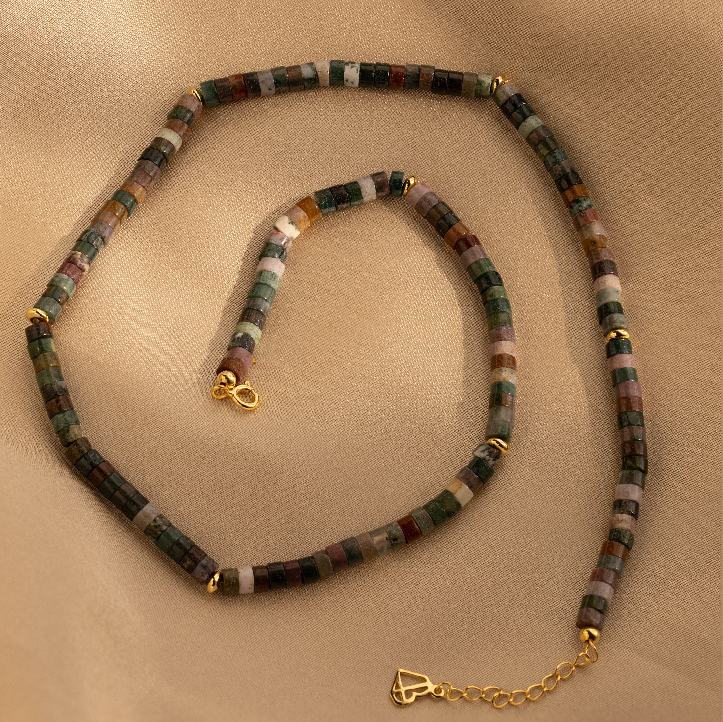 The Moss Agate Beaded Necklace showcases mixed color moss agate beads and gold clasps for earthy elegance, displayed on a beige fabric backdrop.