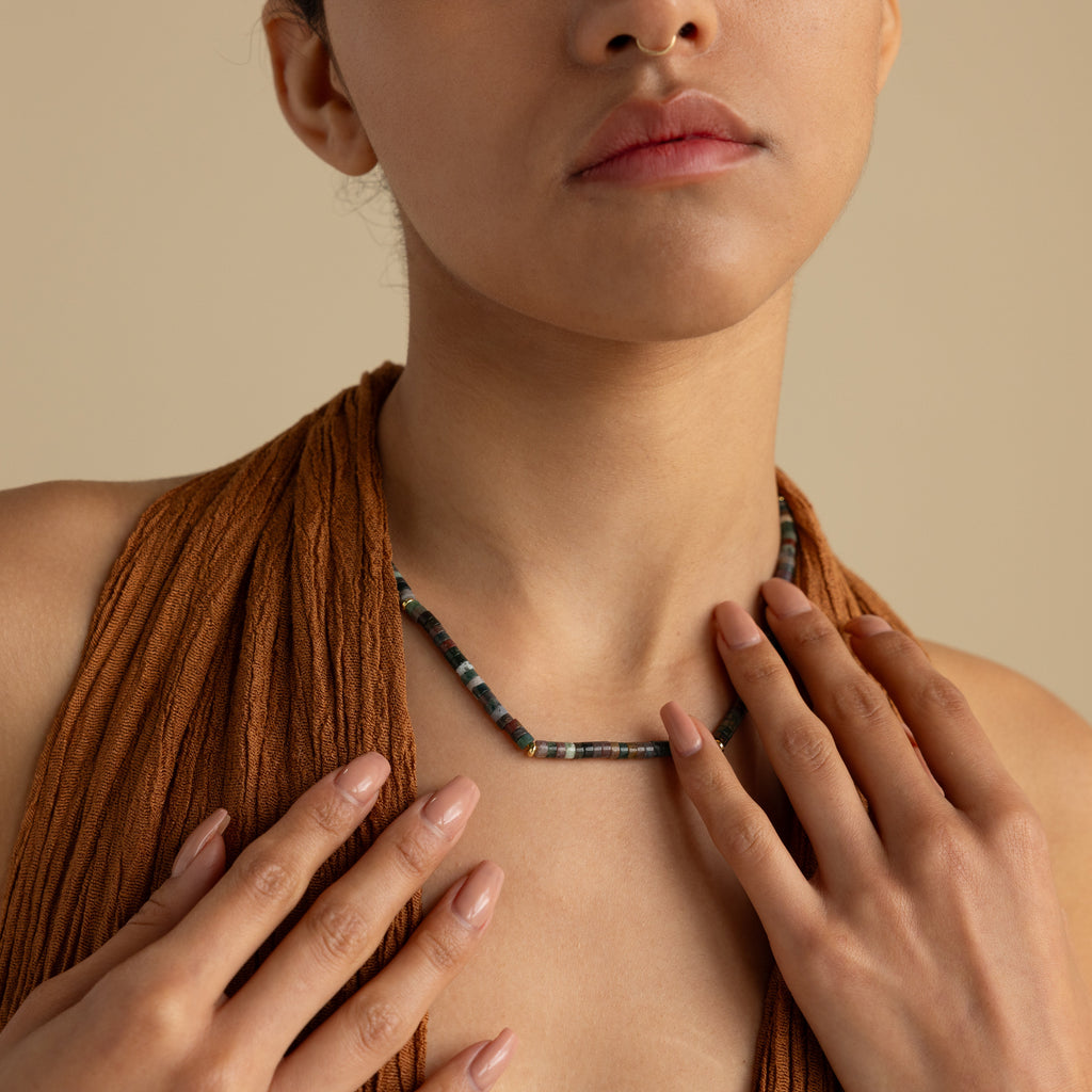 A woman in a textured brown top exudes earthy elegance as she touches her collarbone, showcasing the Moss Agate Beaded Necklace.