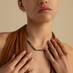 A woman in a textured brown top exudes earthy elegance as she touches her collarbone, showcasing the Moss Agate Beaded Necklace.