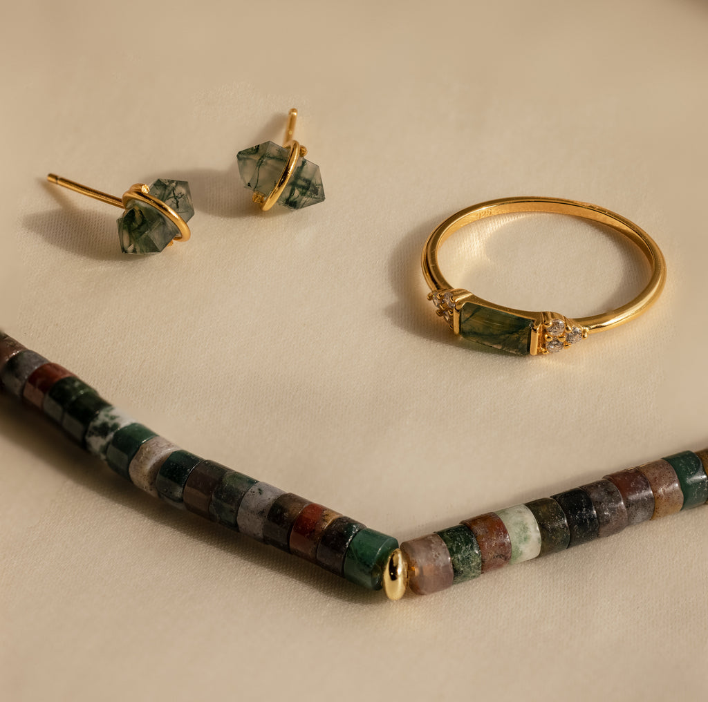 Gold ring and earrings with green stones are set beside a Moss Agate Beaded Necklace, featuring mixed color beads, all beautifully arranged on a beige surface for an earthy, elegant look.