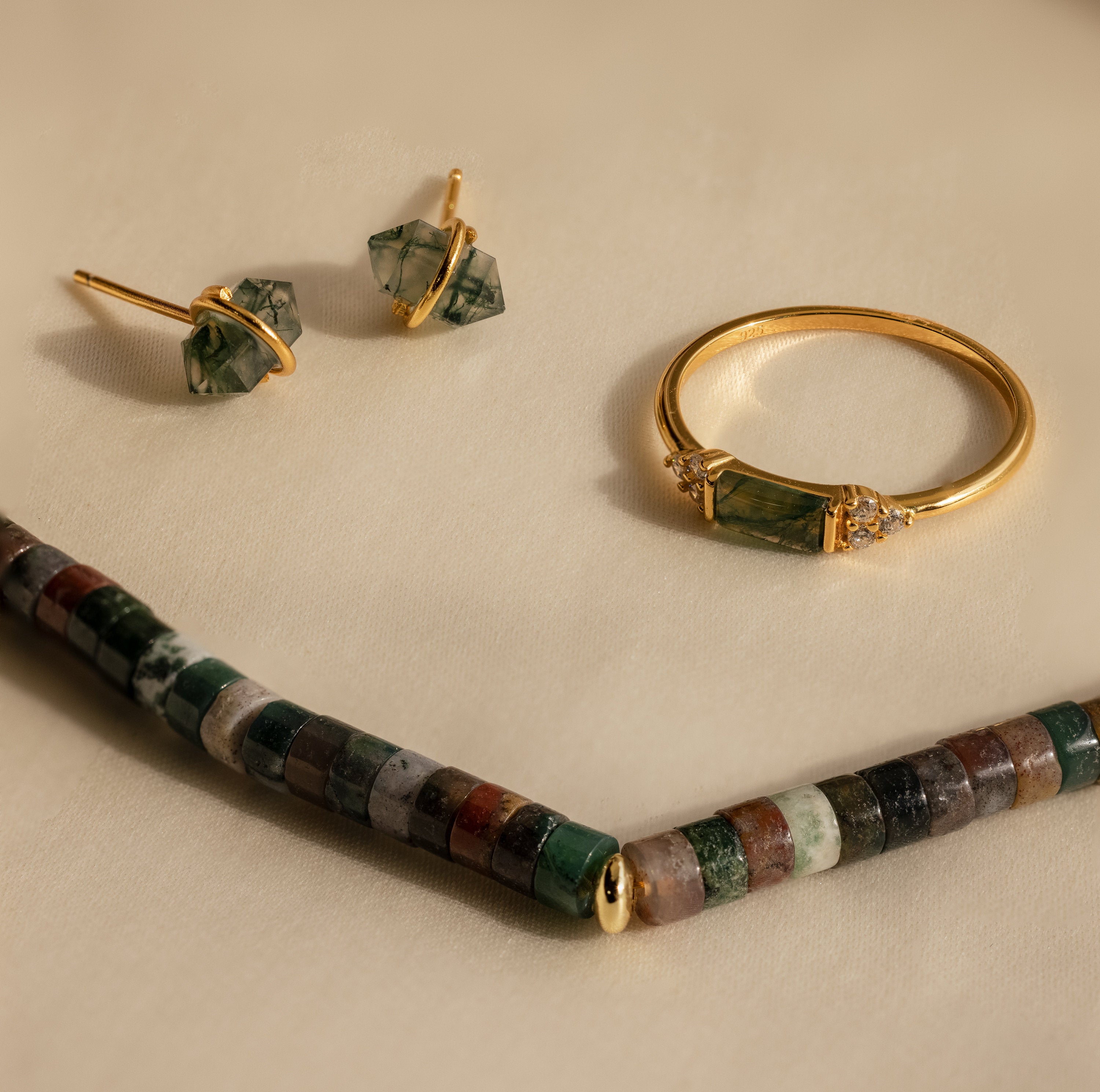 Gold ring and earrings with green stones are set beside a Moss Agate Beaded Necklace, featuring mixed color beads, all beautifully arranged on a beige surface for an earthy, elegant look.