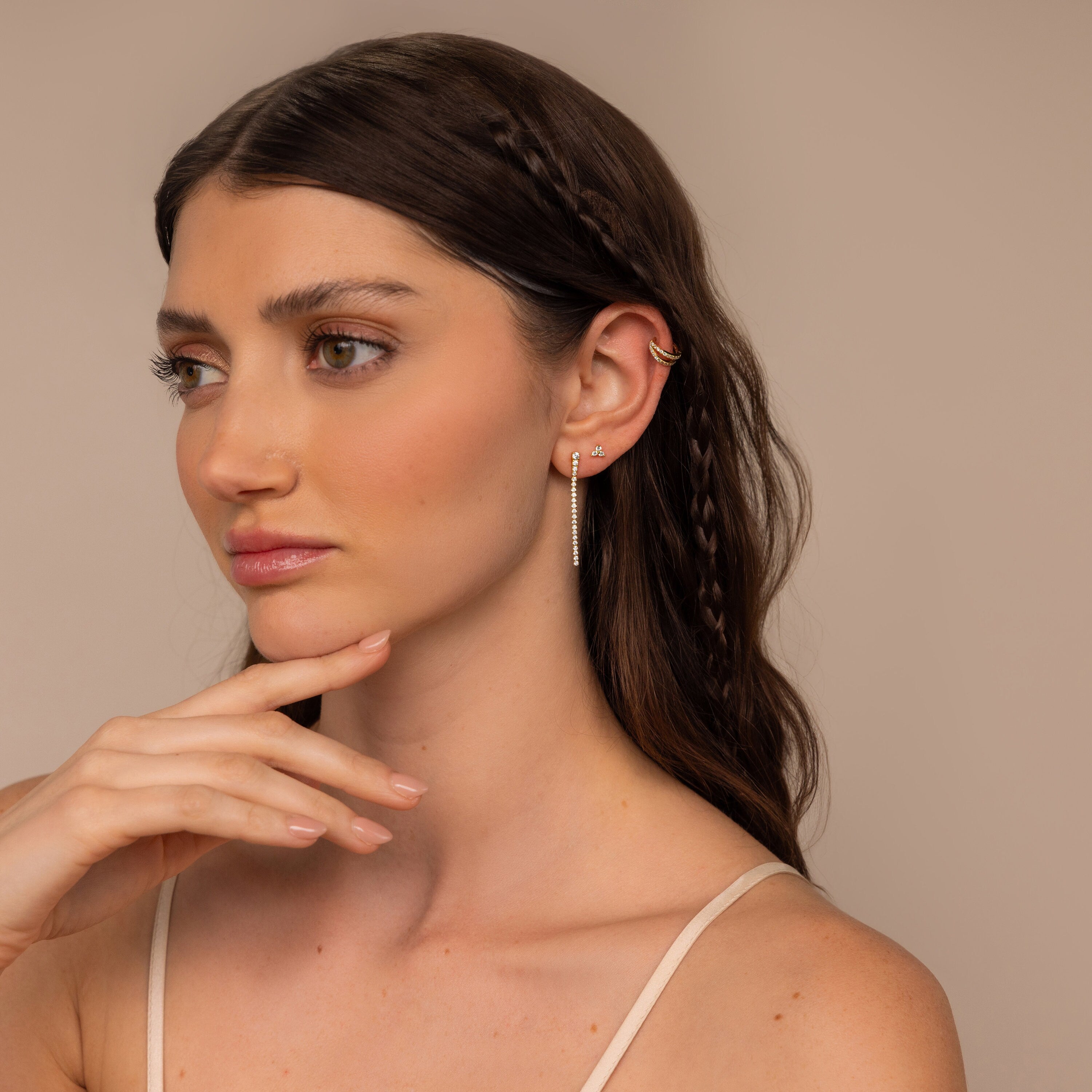 Woman with long brown hair and braided strands gazes thoughtfully to the side, showcasing elegant Diamond Tennis Dangle Earrings.