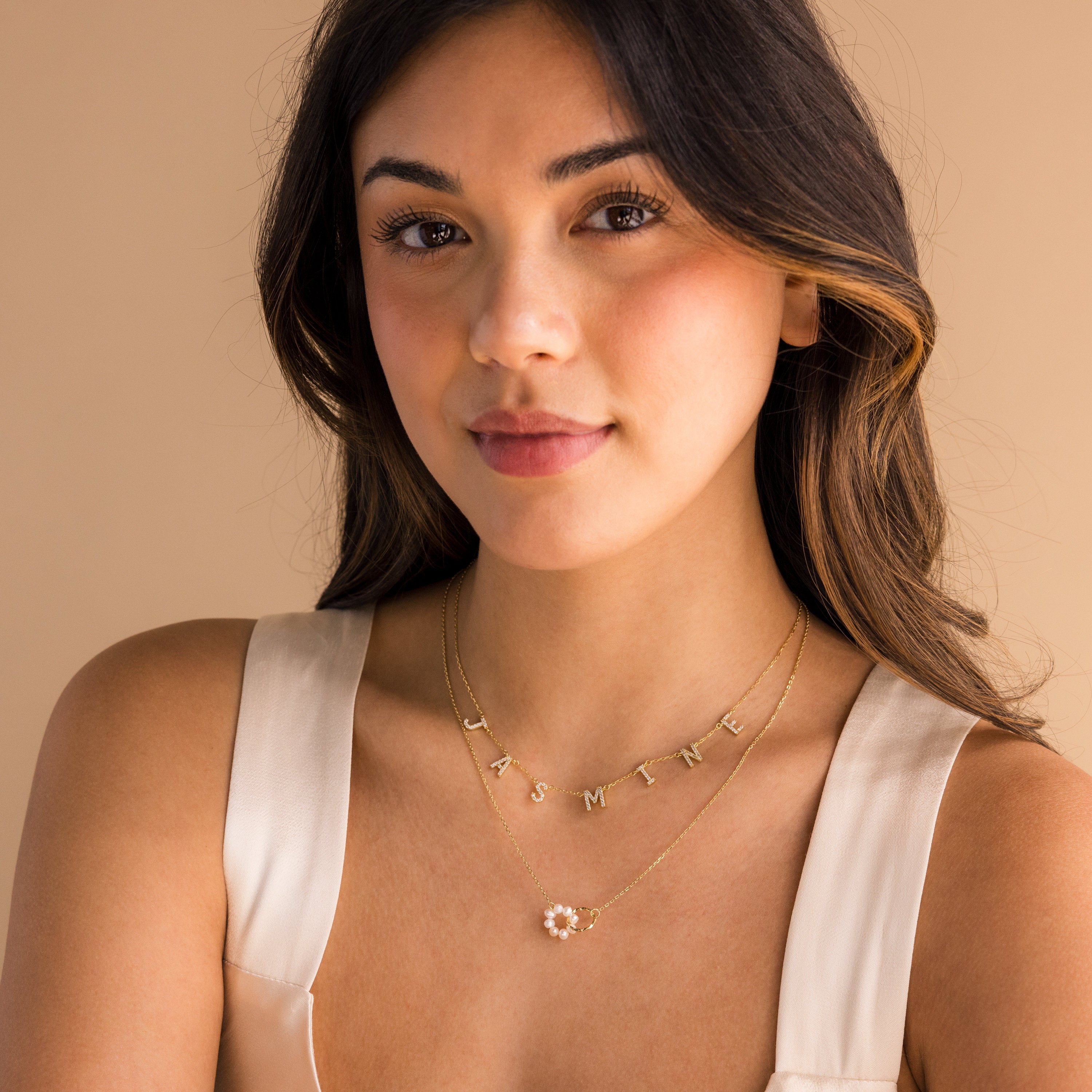 A woman with wavy hair in a cream top layers gold necklaces—one reading JASMINE, another with a flower pendant—and finishes her look with the elegant Interlocking Pearl Necklace.