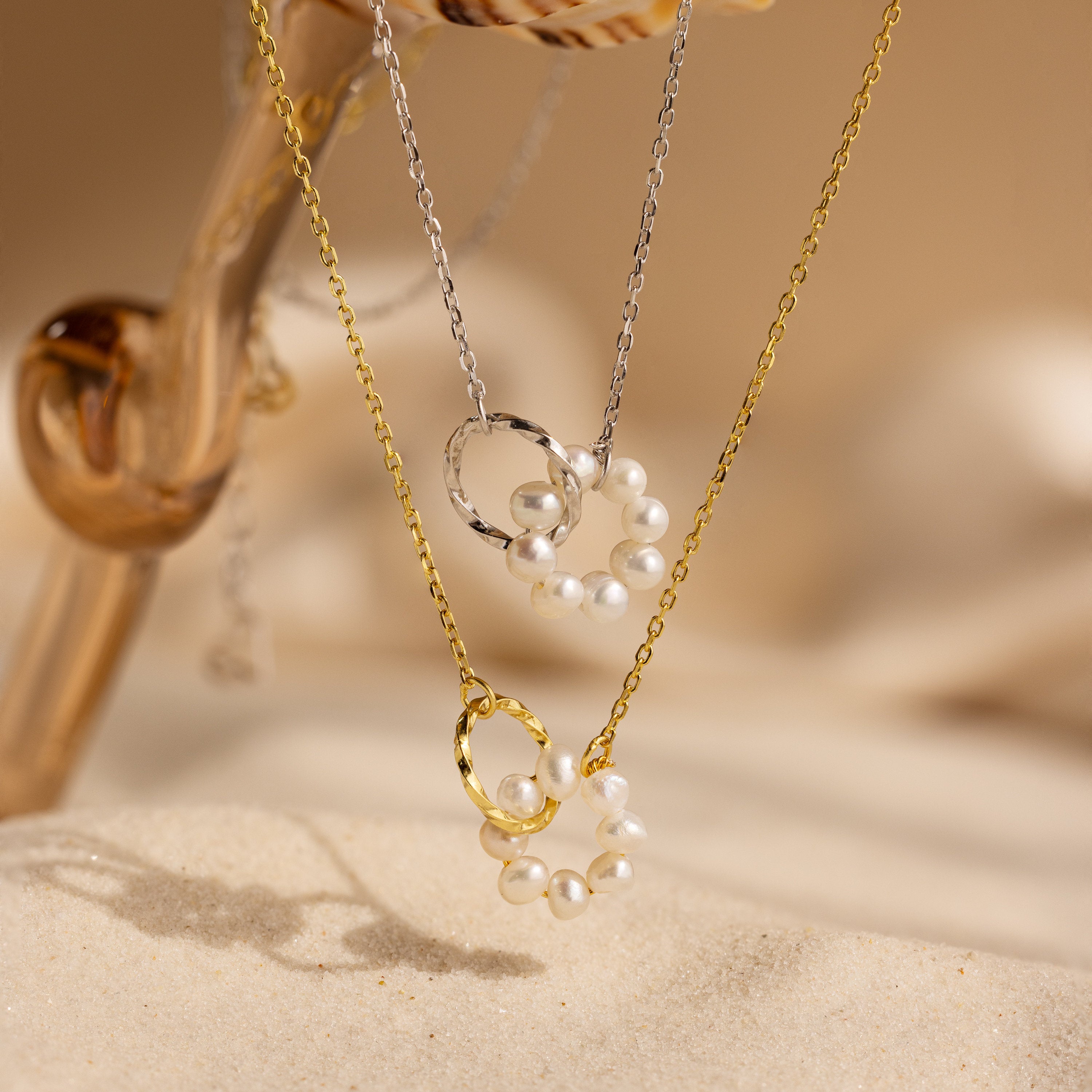 The Interlocking Pearl Necklace features two circular pearl bead pendants—one on a gold chain, one on a silver chain—displayed elegantly against a sandy backdrop.