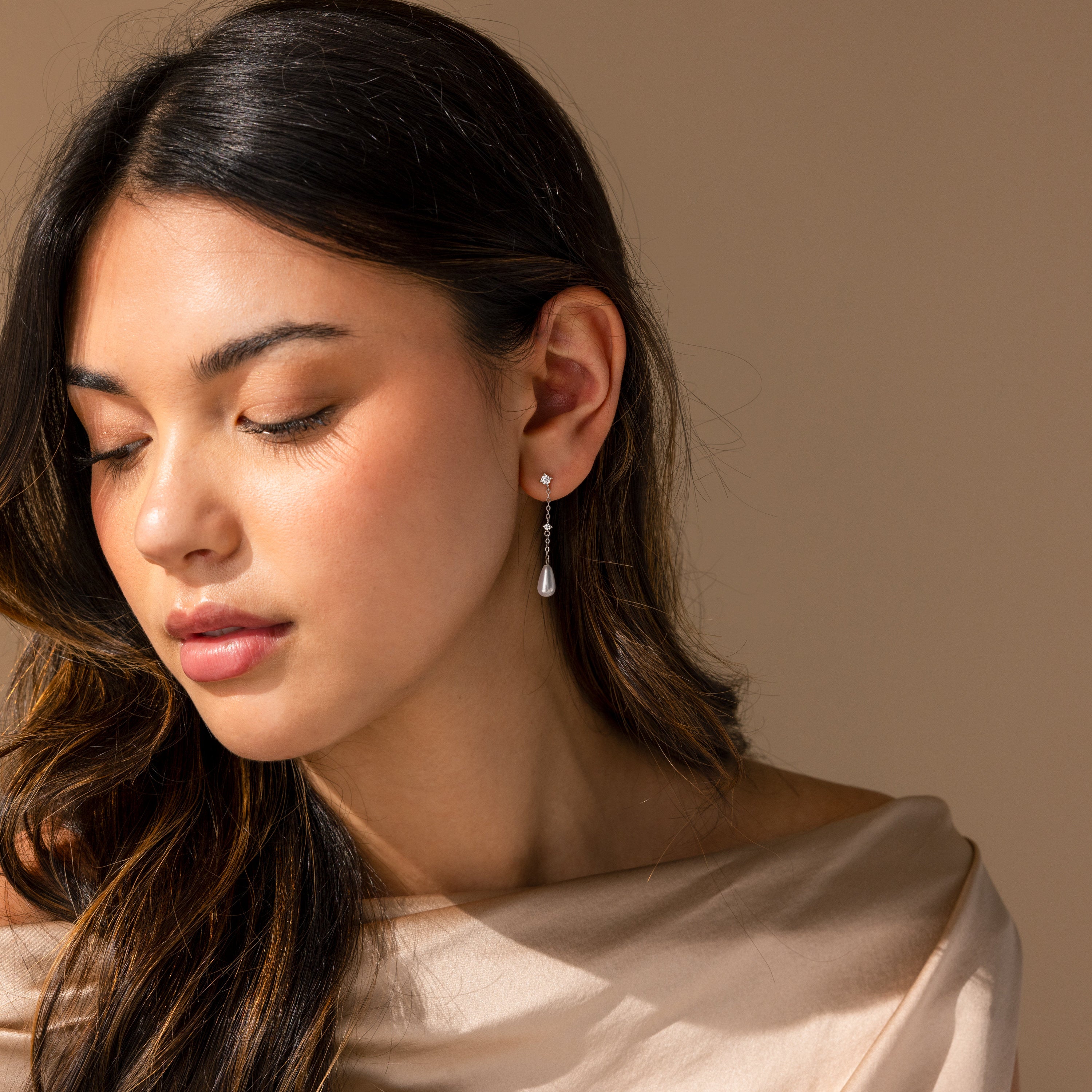 A woman with long hair wearing a satin top and Diamond Pearl Chain Earrings looks down in soft lighting.