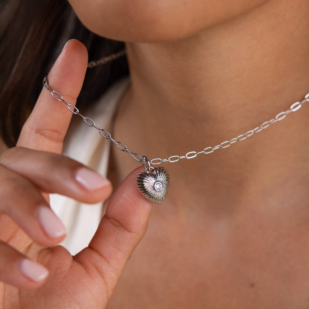 A woman holds the Diamond Ribbed Heart Necklace close to her neck, highlighting its vintage-inspired jewelry charm.