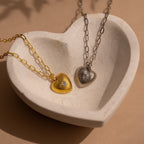 The Diamond Ribbed Heart Necklace in gold and a silver heart necklace are beautifully presented in a cream-colored heart-shaped dish, offering a charming display of vintage-inspired jewelry.