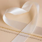 A white satin ribbon forms a heart shape beside two delicate gold and silver chains, featuring the Heart Link Chain Necklace, all displayed on a beige surface.