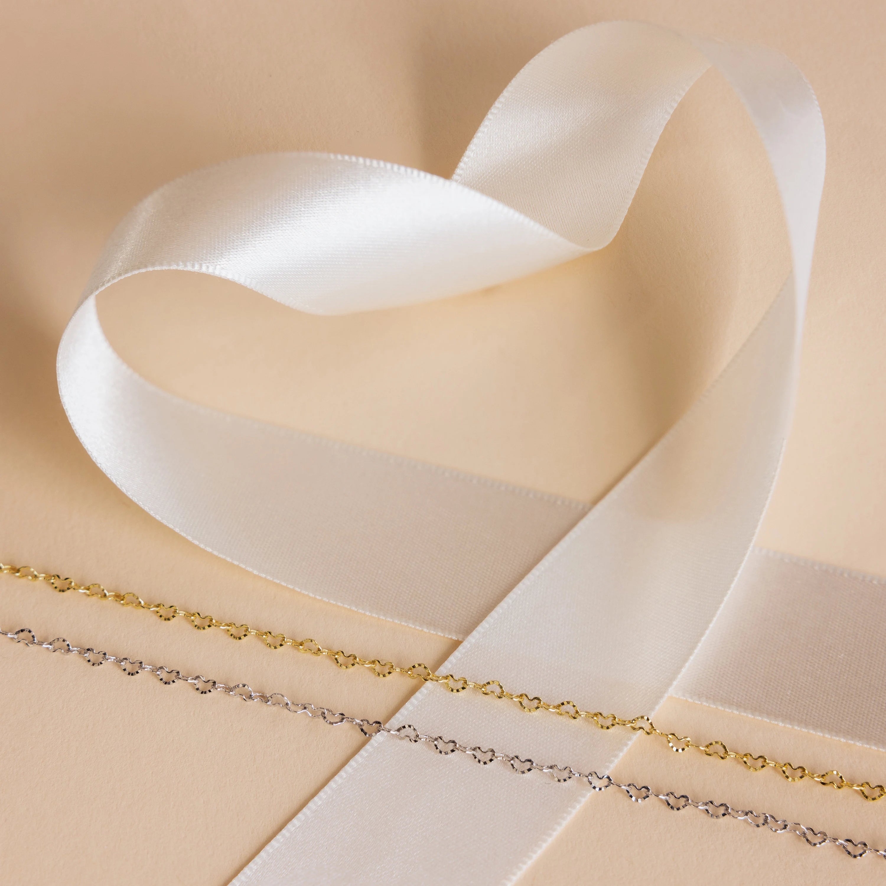 A white satin ribbon forms a heart shape beside two delicate gold and silver chains, featuring the Heart Link Chain Necklace, all displayed on a beige surface.