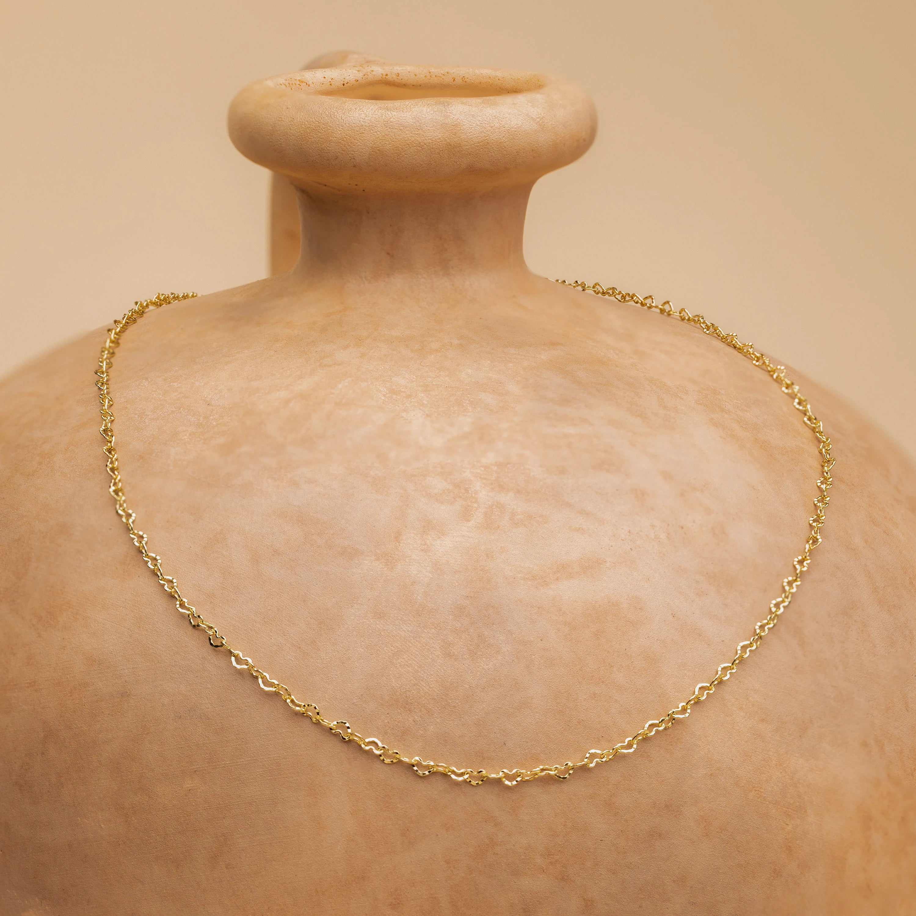 The Heart Link Chain Necklace, featuring interlocking hearts, is draped over a beige ceramic vase for a subtle romantic accent.