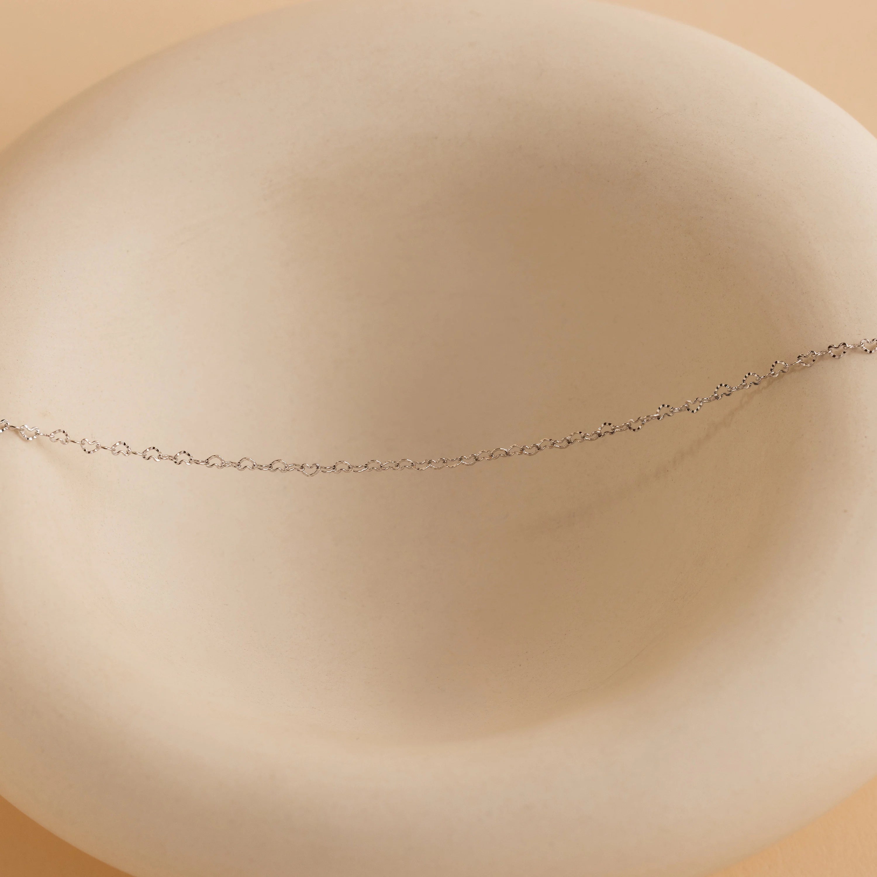The Heart Link Chain Necklace, featuring interlocking hearts on a delicate silver chain, is displayed on a smooth, round, off-white ceramic dish.