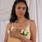 A woman in a white dress stands before a light fabric backdrop, holding a straw bag of white tulips. Her look is finished with the Heart Link Chain Necklace, adding a subtle romantic touch.