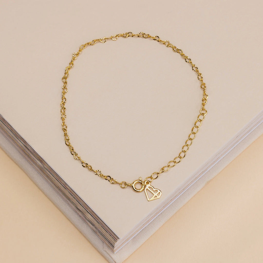 The Heart Link Chain Bracelet, featuring a delicate heart charm, rests on a closed beige book—ideal for anyone who loves romantic jewelry.