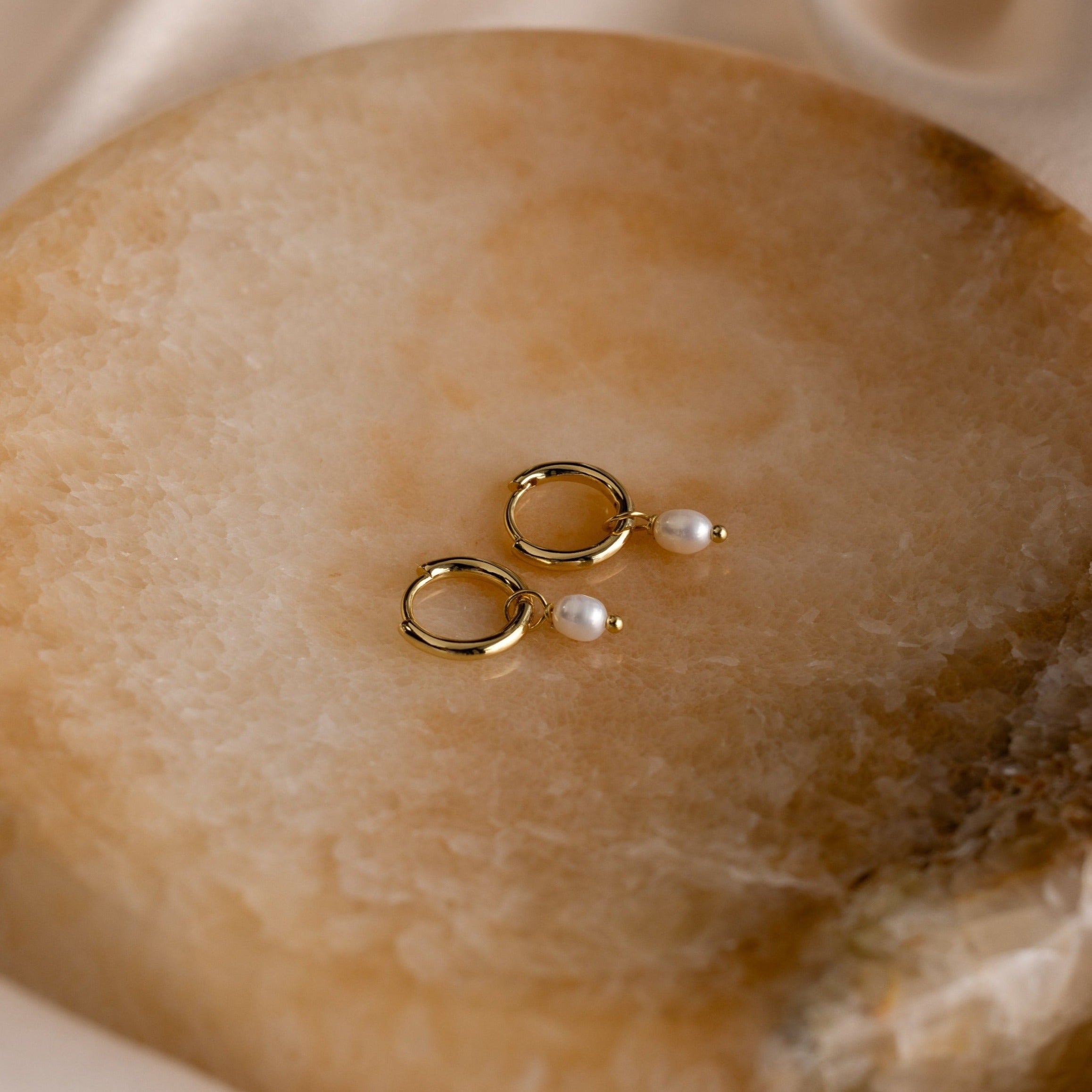 Close-up of Dainty Gold Pearl Huggie Earrings laying on a brown marble dish. 