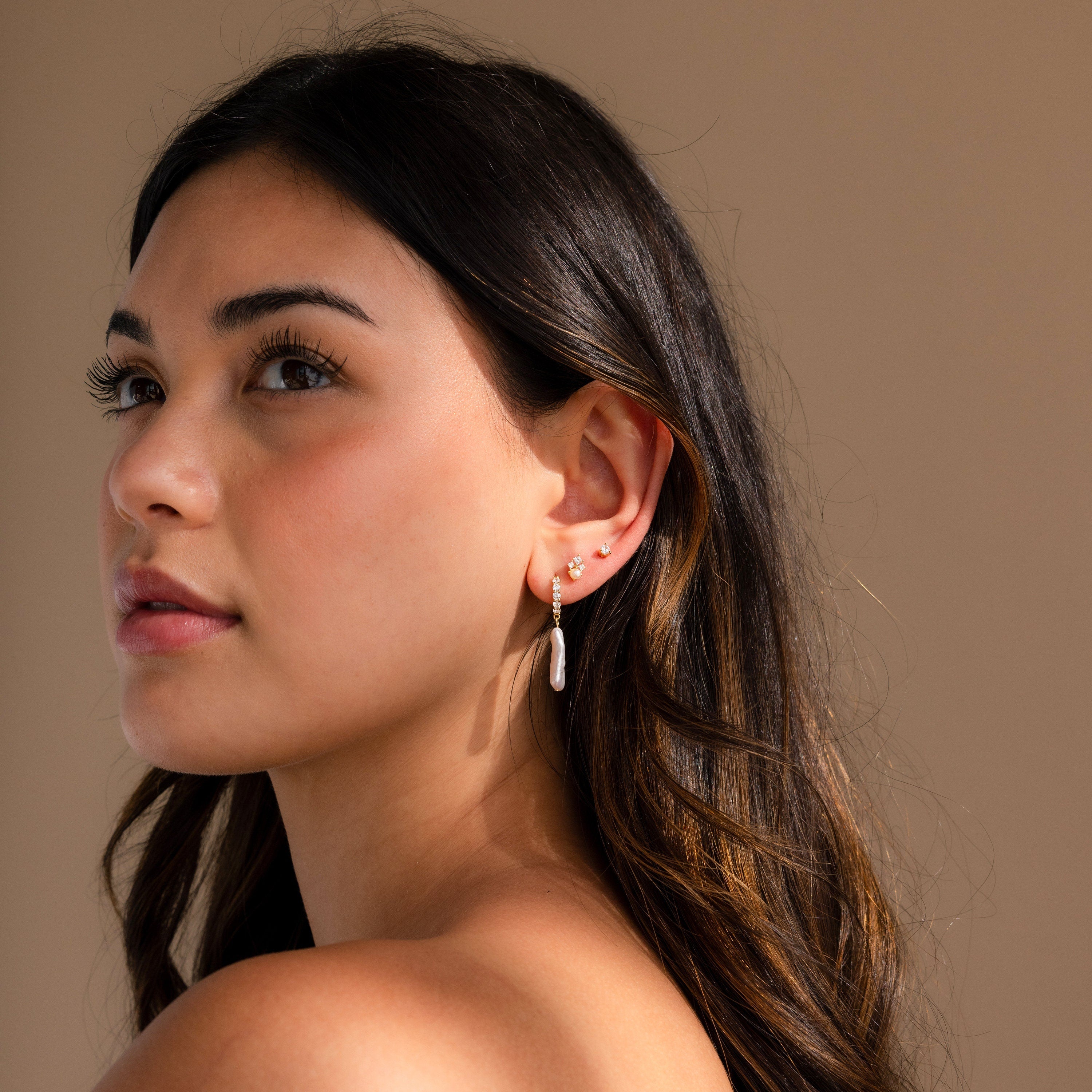 Woman with long dark hair looks up against a neutral background, wearing multiple gold hoop earrings and Baroque Pearl Pave Huggies.