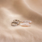 The Baroque Pearl Pave Huggies, featuring silver hoops adorned with Baroque pearls and small crystals, rest on a soft beige fabric background.