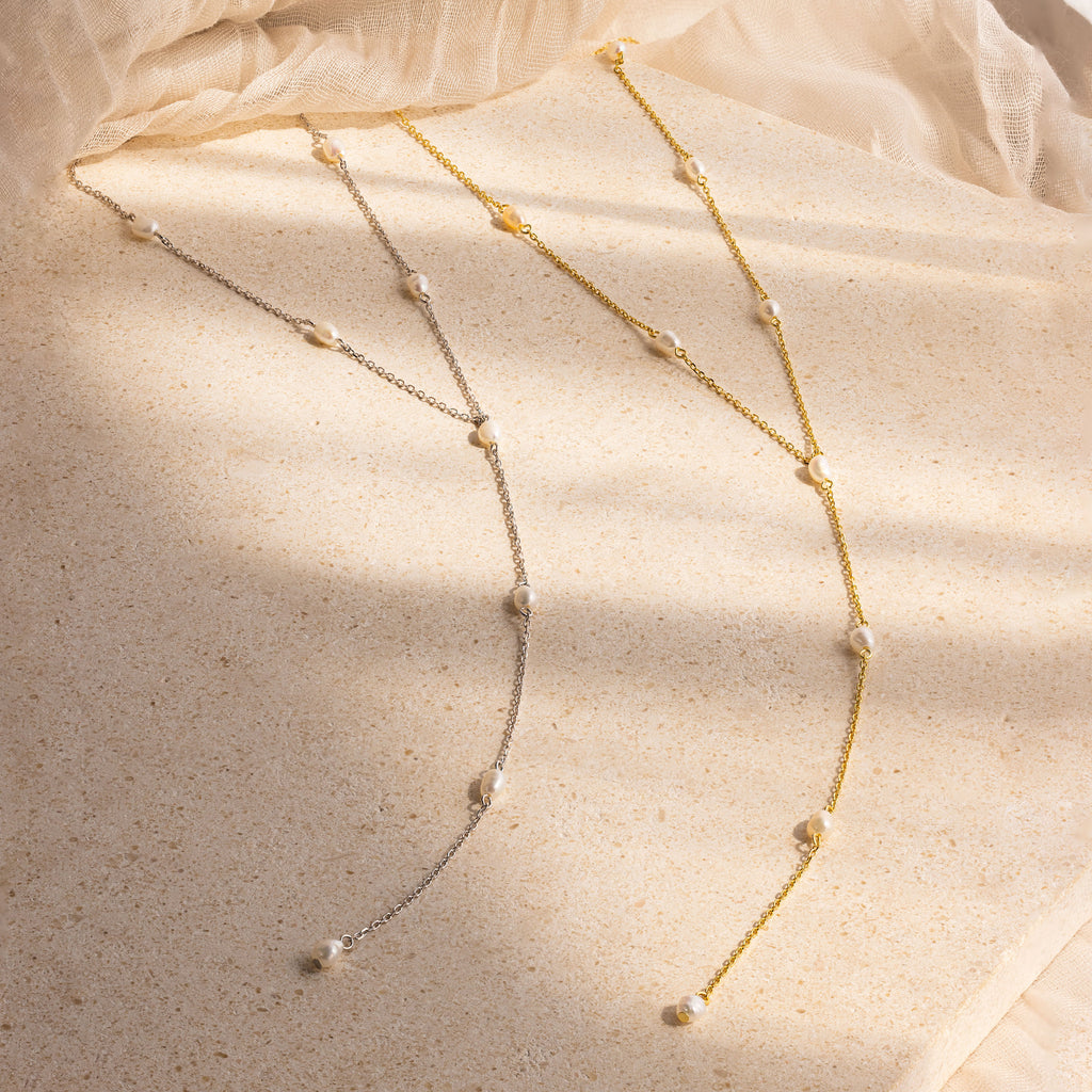 The Pearl Station Lariat Necklace in gold and silver, each adorned with delicate pearls and subtle pearl charms, displayed on a textured beige surface.