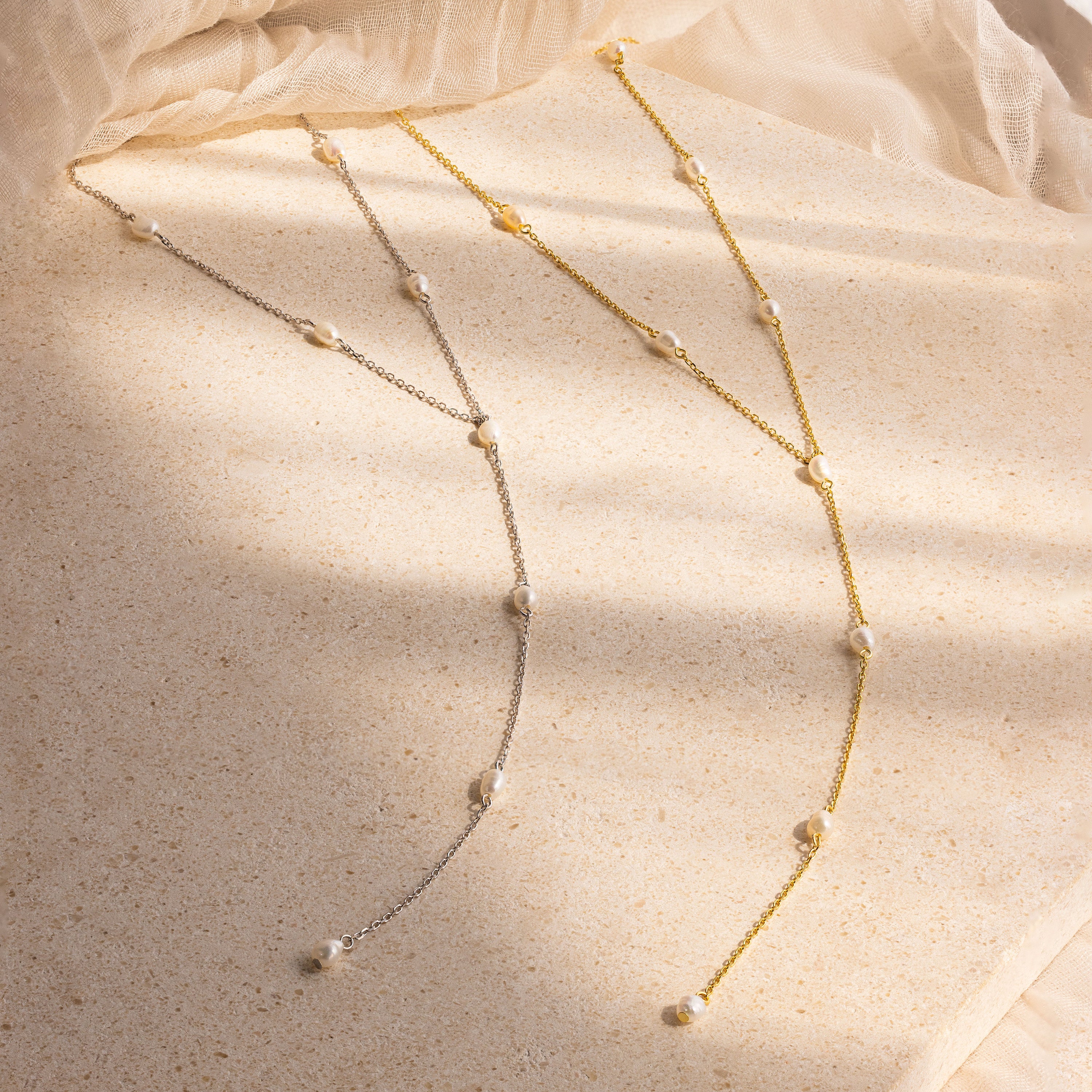 The Pearl Station Lariat Necklace in gold and silver, each adorned with delicate pearls and subtle pearl charms, displayed on a textured beige surface.