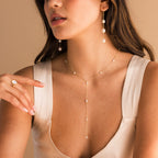 A woman wearing the Pearl Station Lariat Necklace, matched with earrings and a ring, styled with a cream sleeveless dress against a beige background.