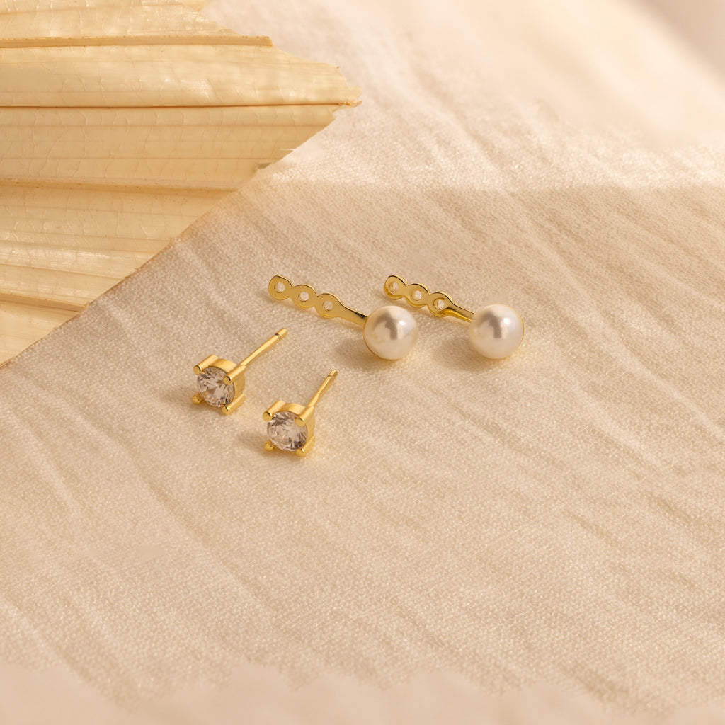 The Diamond Pearl Ear Jackets feature two pairs of gold studs accented with pearls and clear gems, displayed on beige textured fabric—ideal for fans of minimalist jewelry.