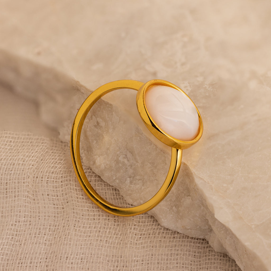 The Mother of Pearl Signet Ring in 18K Gold, featuring a round white stone, rests on a textured beige surface.