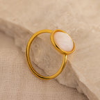 The Mother of Pearl Signet Ring in 18K Gold, featuring a round white stone, rests on a textured beige surface.