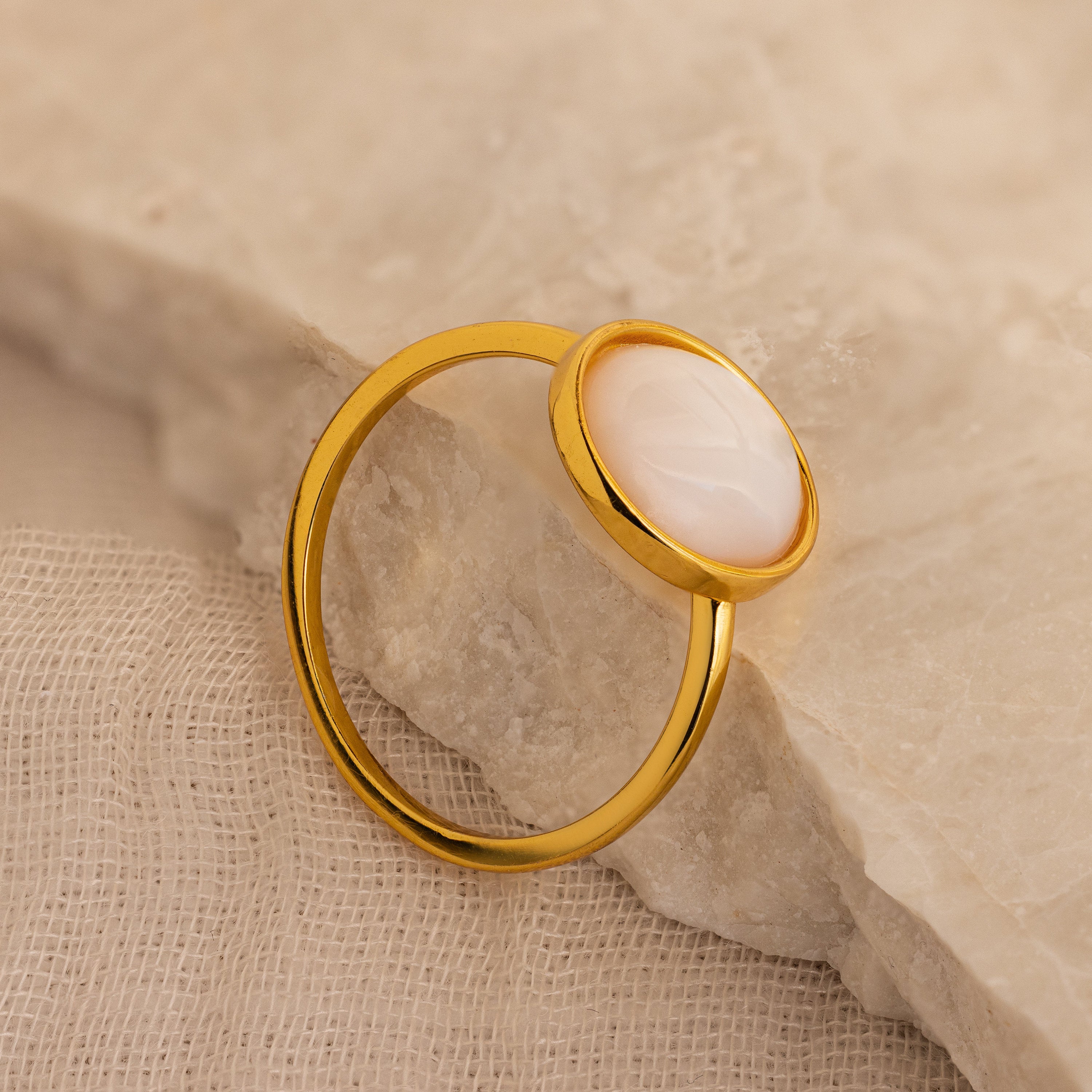 The Mother of Pearl Signet Ring in 18K Gold, featuring a round white stone, rests on a textured beige surface.