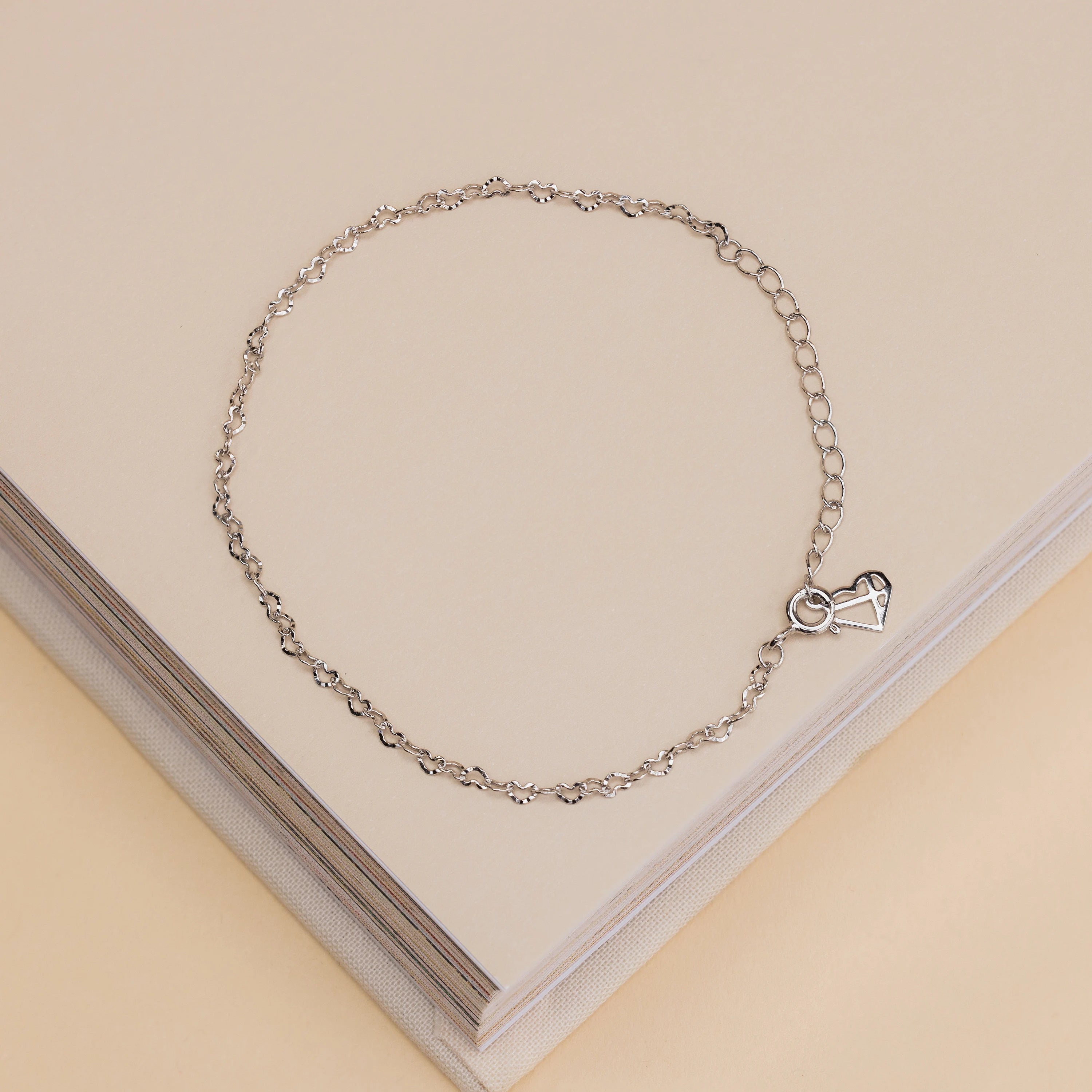The Heart Link Chain Bracelet, a delicate silver piece with a heart-shaped lock charm, is elegantly displayed on a beige book.