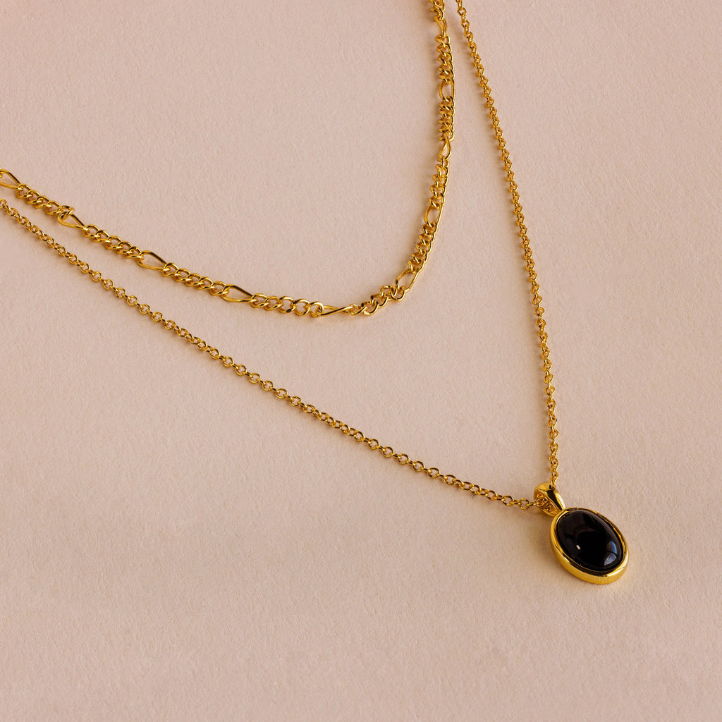 The Onyx Figaro Duo Necklace, featuring a classic Figaro Chain and an elegant onyx pendant, is displayed on a light beige surface, highlighting its refined, stylish design.