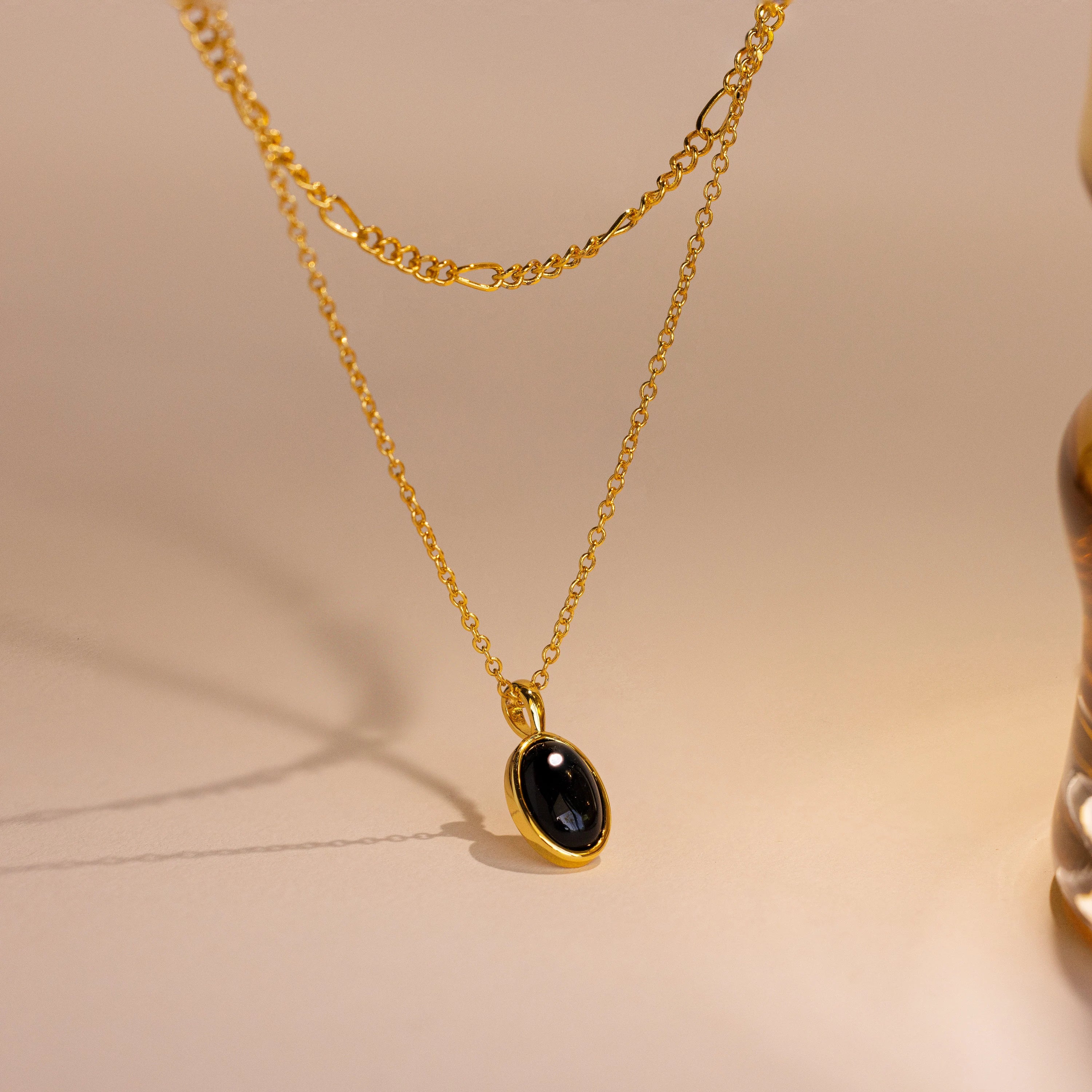 The Onyx Figaro Duo Necklace features a striking onyx pendant on an elegant Figaro chain, displayed against a neutral background with soft lighting to highlight its sophisticated design.