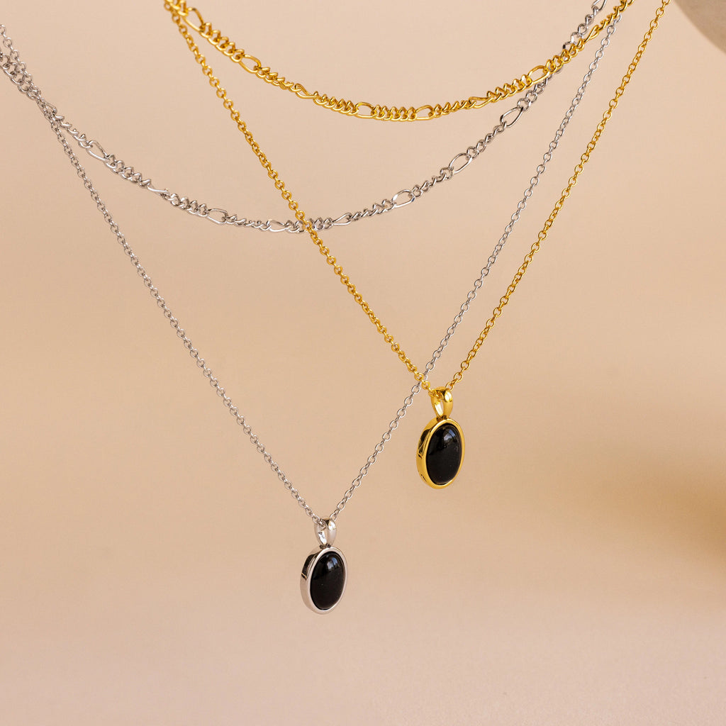 The Onyx Figaro Duo Necklace set features four layered chains—two gold, two silver—with sleek black onyx pendants and classic Figaro links, styled together and displayed on a beige background.