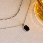 The Onyx Figaro Duo Necklace, featuring a black oval pendant, is displayed on a cream surface beside a yellow glass object.