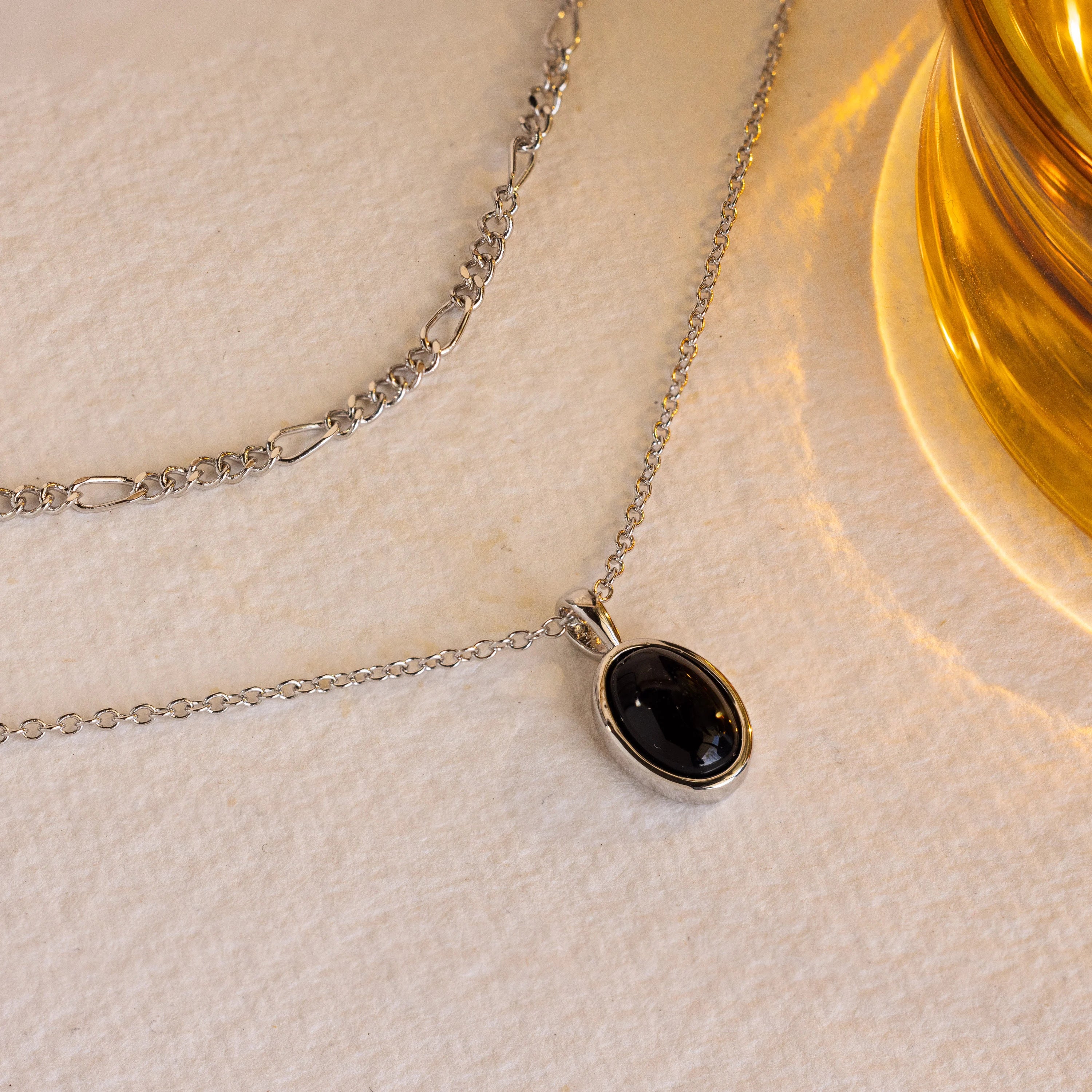 The Onyx Figaro Duo Necklace, featuring a black oval pendant, is displayed on a cream surface beside a yellow glass object.