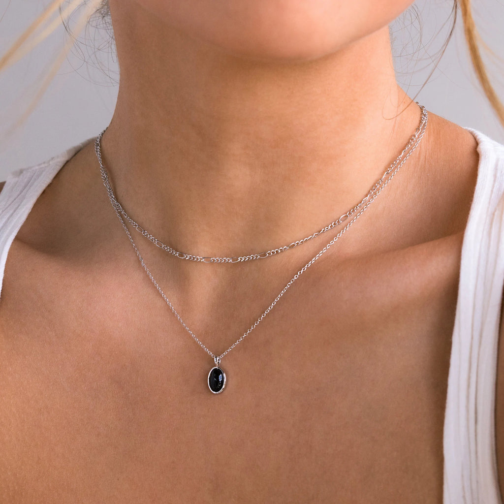 A woman wears the Onyx Figaro Duo Necklace—two silver Figaro chains, one with a bold black onyx pendant—over a white tank top.