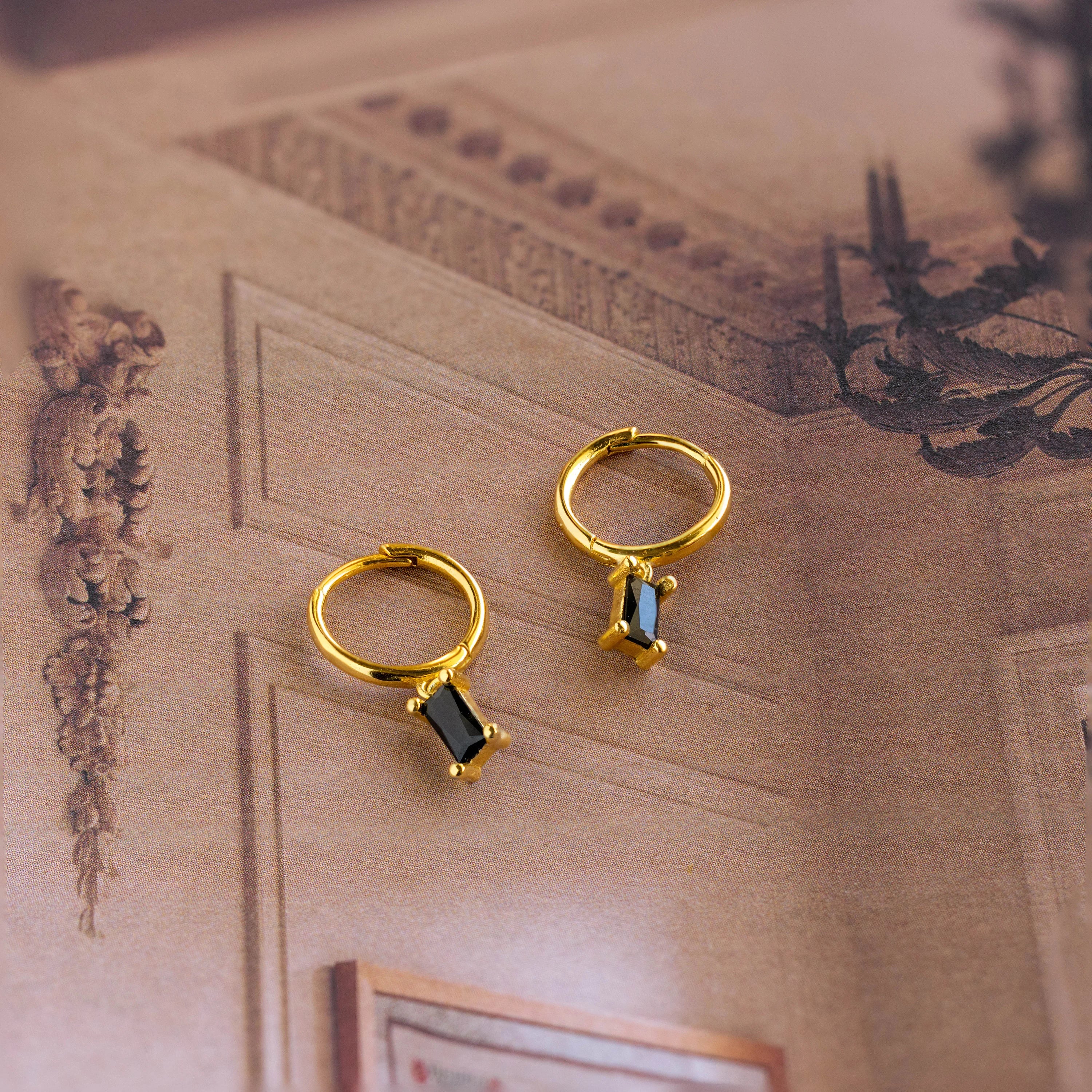 Two Onyx Baguette Drop Huggies—gold hoops featuring black rectangular stones—are showcased on a vintage-inspired decorative background.